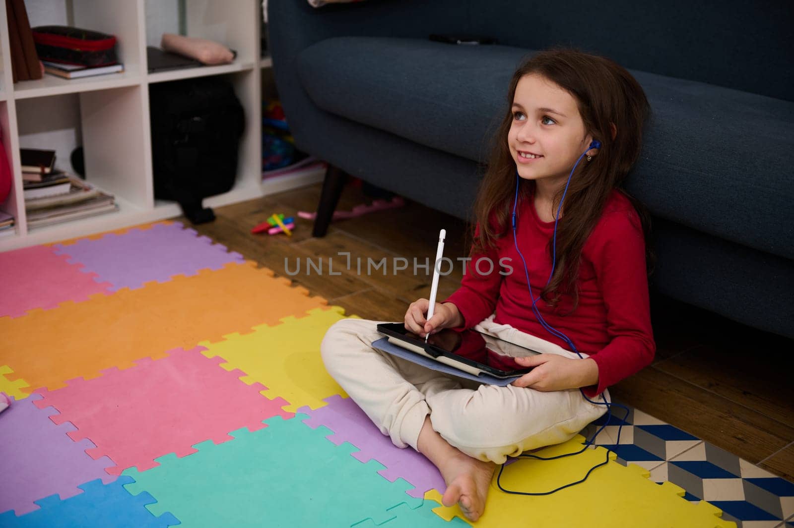 Smiling child girl doing homework on tablet, studying online sitting on multicolored puzzle carpet at home. Distance learning and kids education concept. Copy ad space on black mockup touch screen by artgf