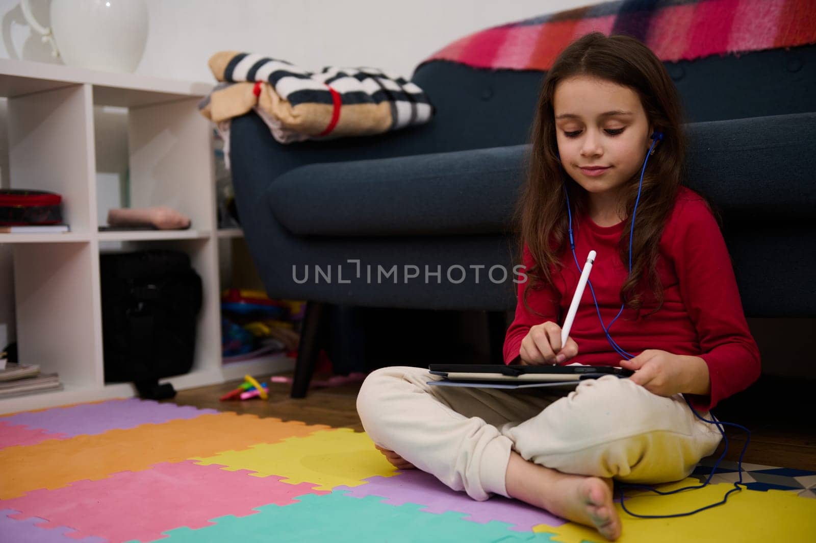 Elementary age student girl in earphones, listening to online class, watching webinar on digital tablet, sitting on multi colored puzzle carpet at home interior. People. Education. Digital technology