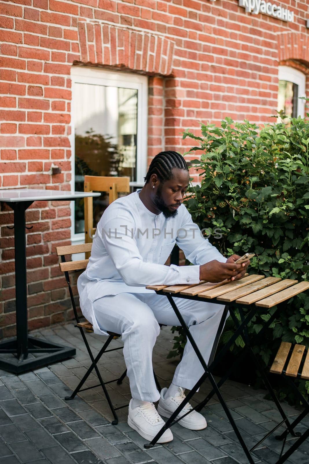 African american man in stylish ethnic dashiki wear viewing photos in social networks on mobile enjoying free time outdoors. Dark-skinned man watching funny video on smartphone and social networks. Blog and blogger scrolling content in Internet by Satura86