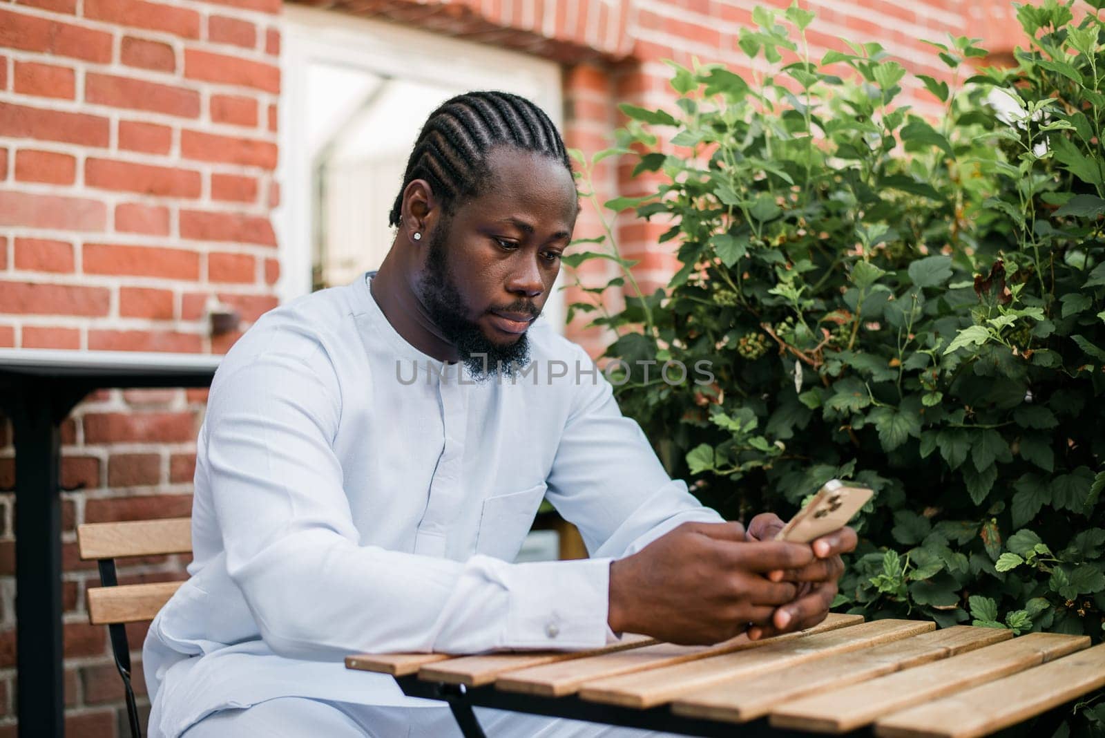 African american man in stylish ethnic dashiki wear viewing photos in social networks on mobile enjoying free time outdoors. Dark-skinned man watching funny video on smartphone and social networks. Blog and blogger scrolling content in Internet by Satura86