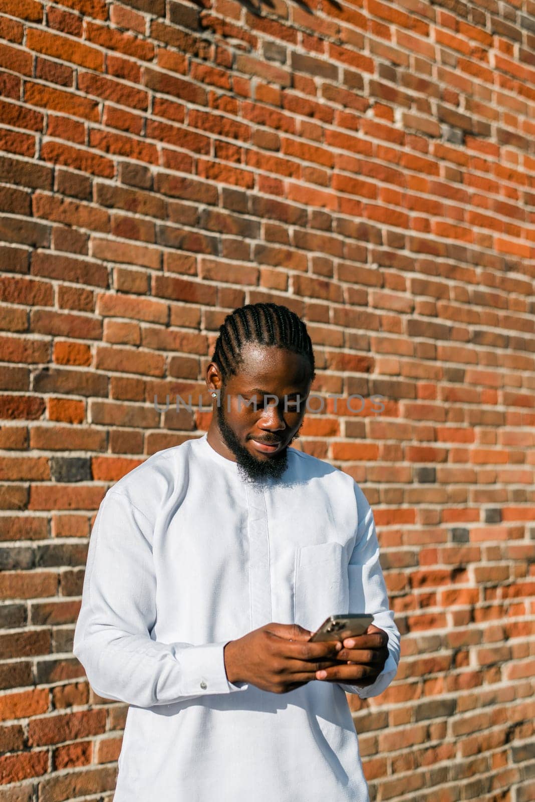 African american man in stylish ethnic dashiki wear viewing photos in social networks on mobile enjoying free time outdoors. Dark-skinned man watching funny video on smartphone and social networks. Blog and blogger scrolling content in Internet by Satura86
