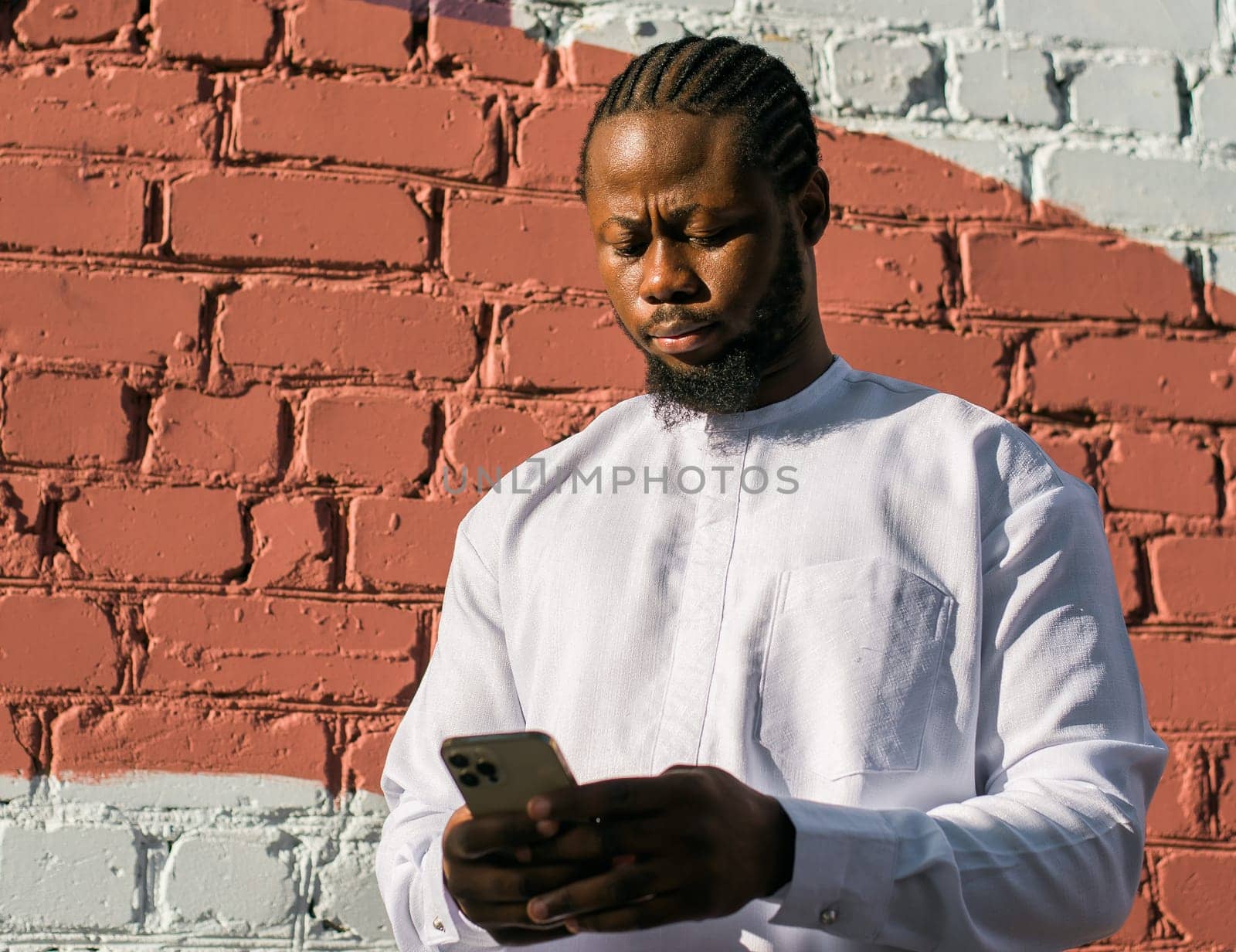 African american man in stylish ethnic dashiki wear viewing photos in social networks on mobile enjoying free time outdoors. Dark-skinned man watching funny video on smartphone and social networks. Blog and blogger scrolling content in Internet.