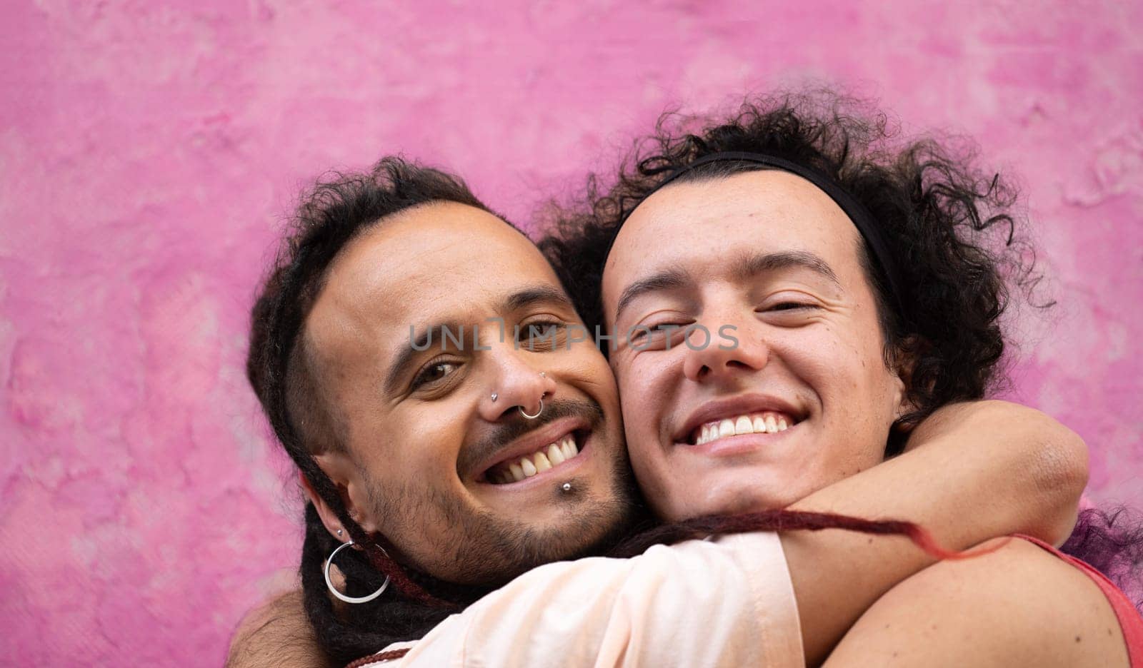Close up headshot of two gay men lovers hug each other with smiles on their faces isolated on pink background.