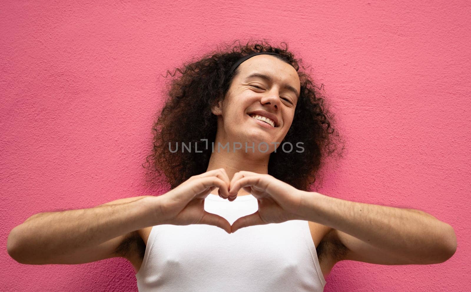 Gay man with curly hair is smiling and making a heart shape with his hands isolated on pink background by papatonic
