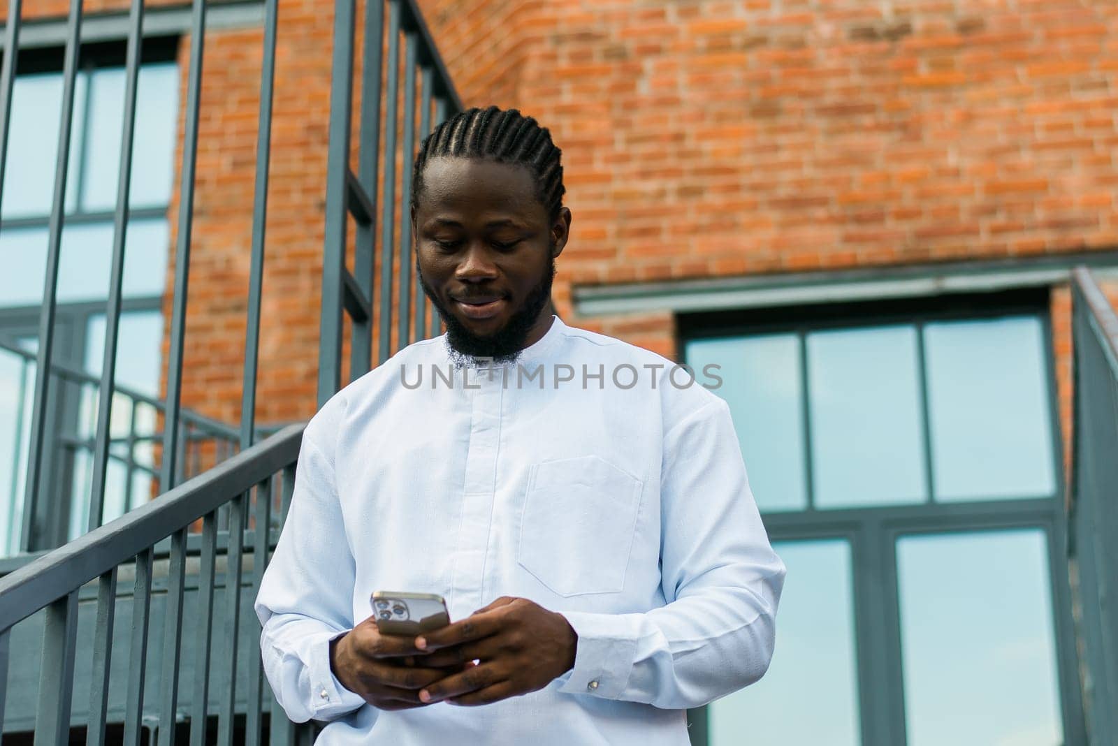 African american man in stylish ethnic dashiki wear viewing photos in social networks on mobile enjoying free time outdoors. Dark-skinned man watching funny video on smartphone and social networks. Blog and blogger scrolling content in Internet.