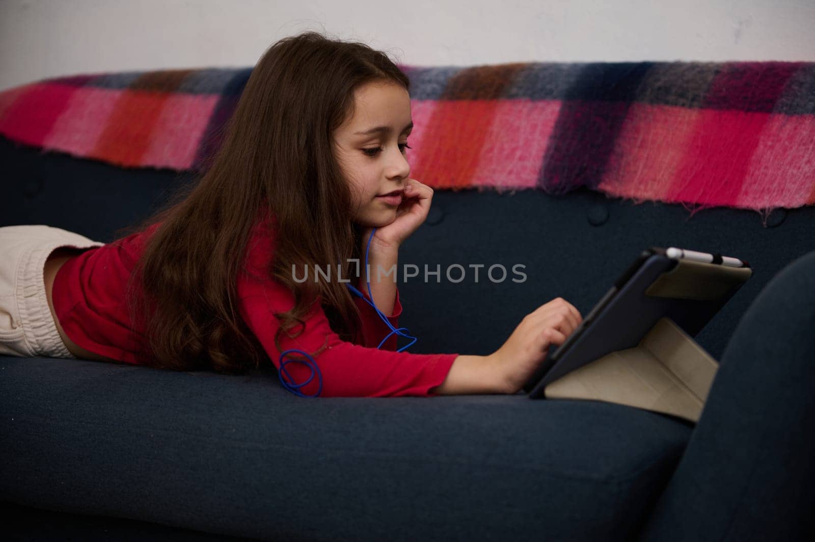 Adorable elementary age kid, cute little kid girl using digital tablet, watching movies while lying on the sofa at cozy home interior. Distance learning. Online education. Digital gadget addiction by artgf