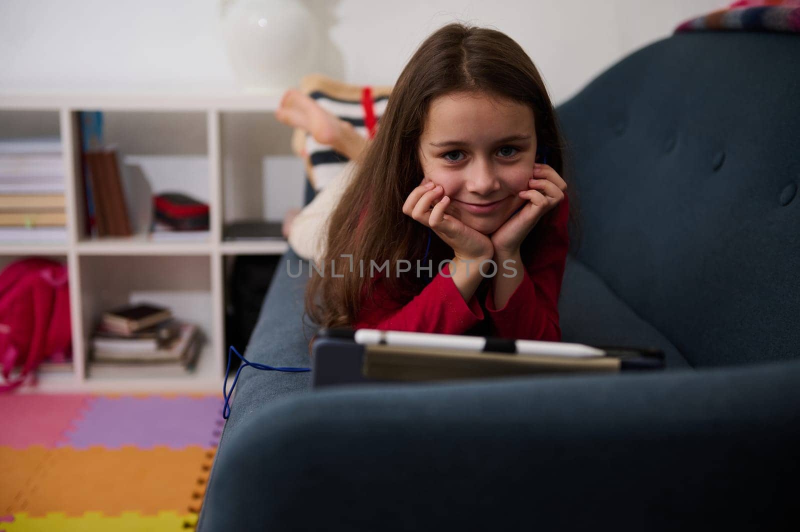 Little kid girl, addicted to digital gadgets, playing online video games while lying on the sofa at home. Lovely school girl watching movies, smiling looking at camera. Happy carefree childhood by artgf