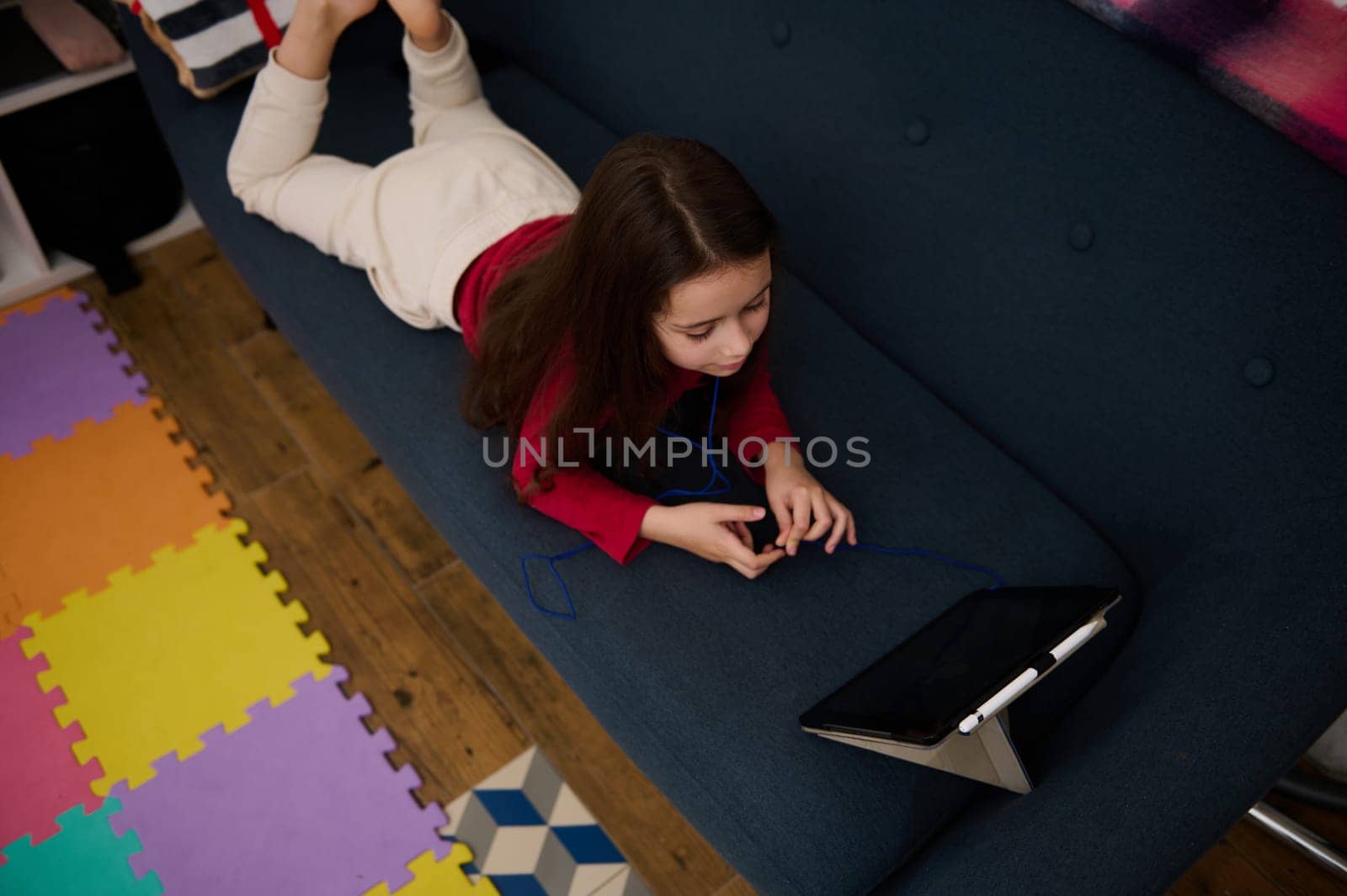 View from above of cute school girl lying on a sofa and using digital tablet at home by artgf