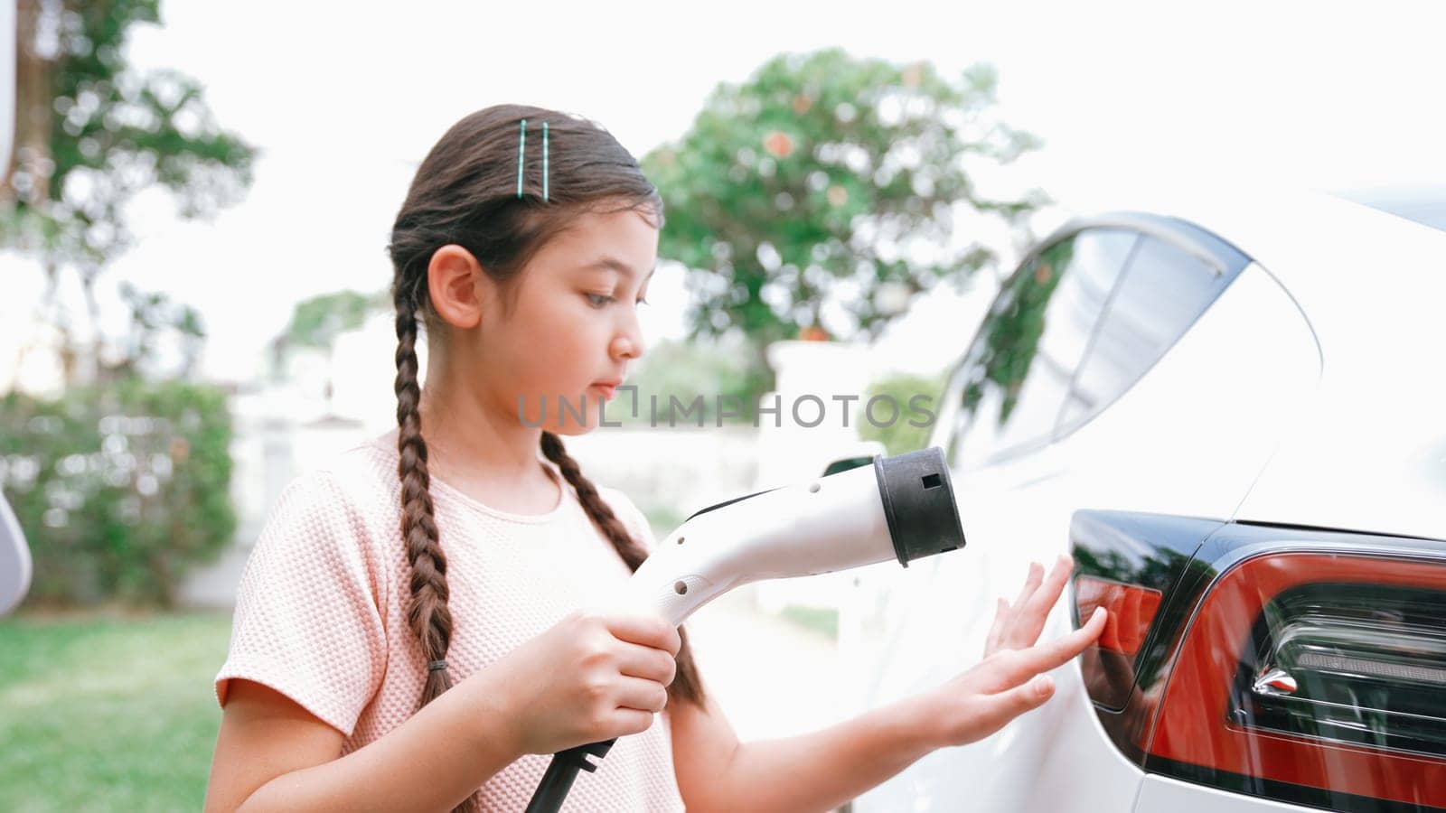 Happy little young girl learn about eco-friendly and energy sustainability as she recharge electric vehicle from home EV charging station. EV car and sustainable future generation concept. Synchronos