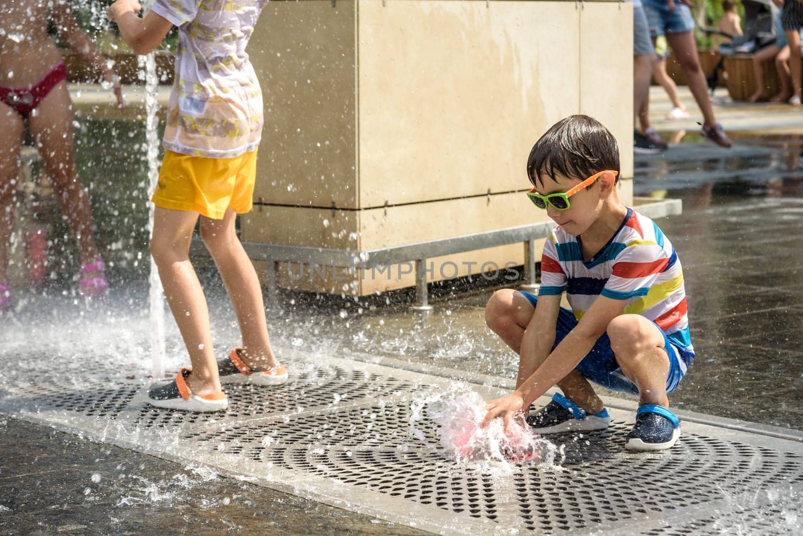 Boy having fun in water fountains. Child playing with a city fountain on hot summer day. Happy kids having fun in fountain. Summer weather. Active leisure, lifestyle and vacation by Kobysh