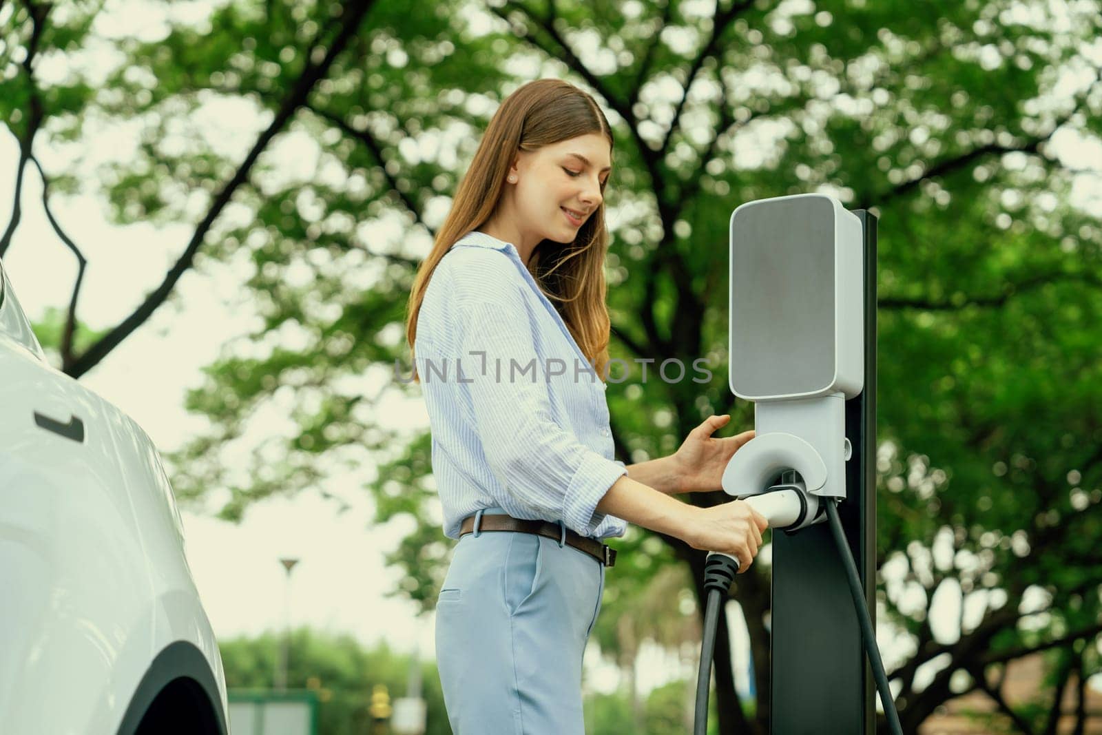 Young woman recharging battery for electric car during road trip travel EV car in natural forest or national park. Eco friendly travel during vacation and holiday. Exalt