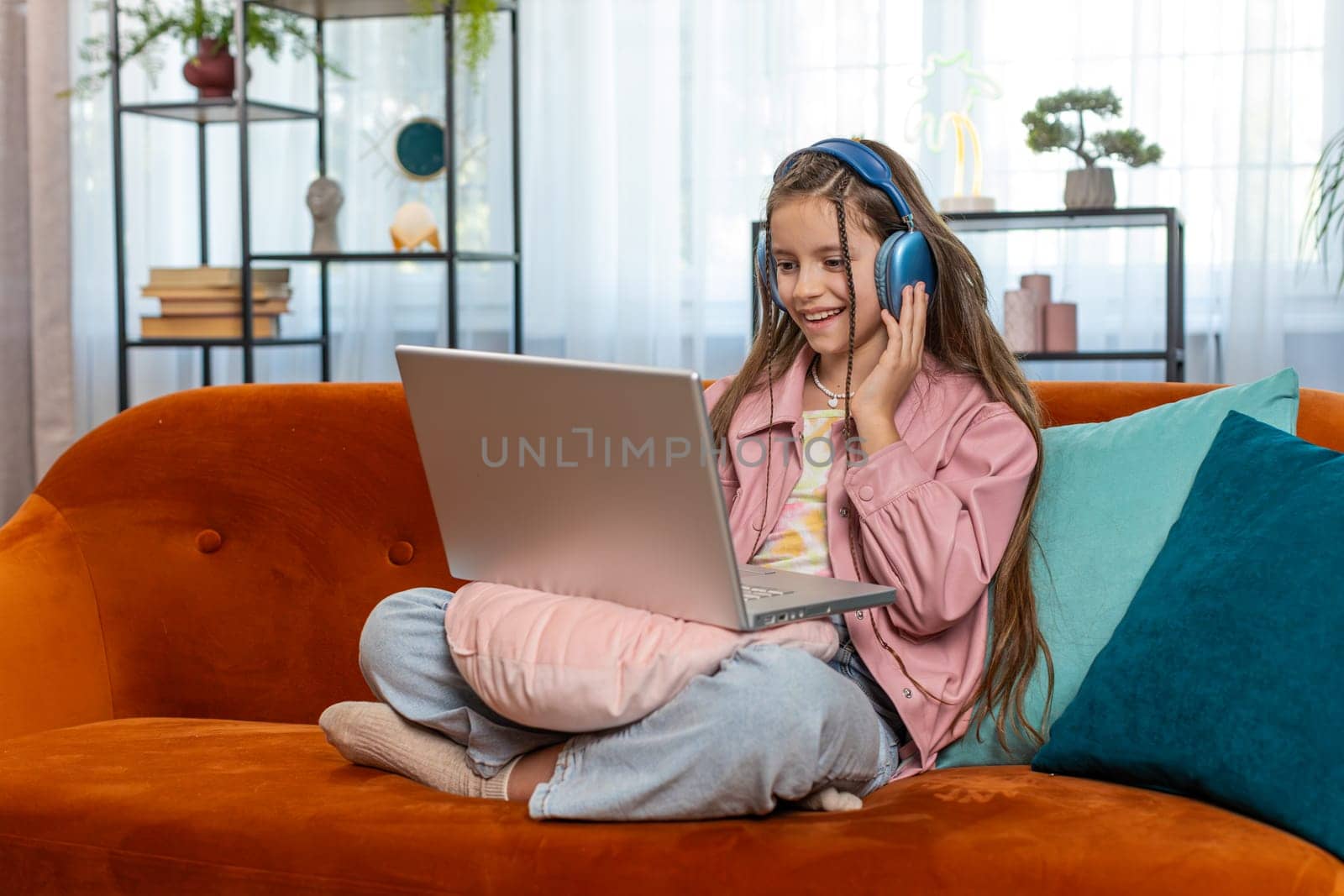 Caucasian preteen girl in headphones using laptop listens to music or lesson, distance learning. Online education. Smiling young child kid relaxing, taking a break at home apartment sitting on couch.