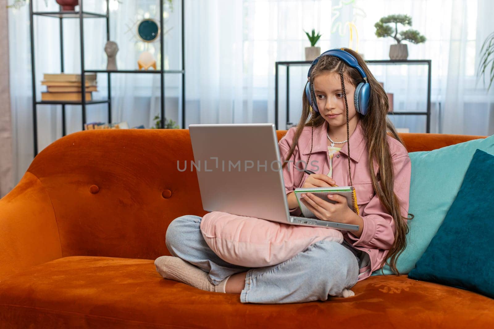 Online education at laptop of little child kid at home virtual class. Remote lesson studying of small school teen girl taking notes and viewing screen. Modern distance e-learning sits on sofa at home.