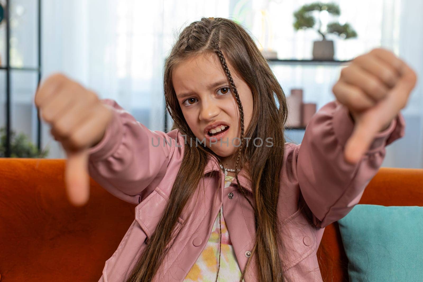 Dislike. Upset girl child showing thumbs down sign gesture, expressing discontent, disapproval, dissatisfied bad work at modern home apartment indoors. Displeased teenager kid in living room on sofa.