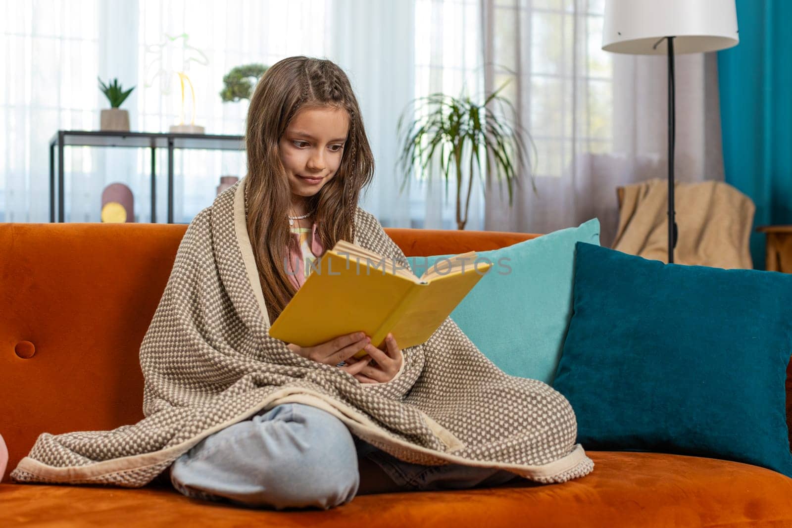 Young Caucasian child wrapped in blanket relaxing reading interesting book turning pages smiling enjoying literature education, taking a rest on couch. Happy girl on couch in living room at home.