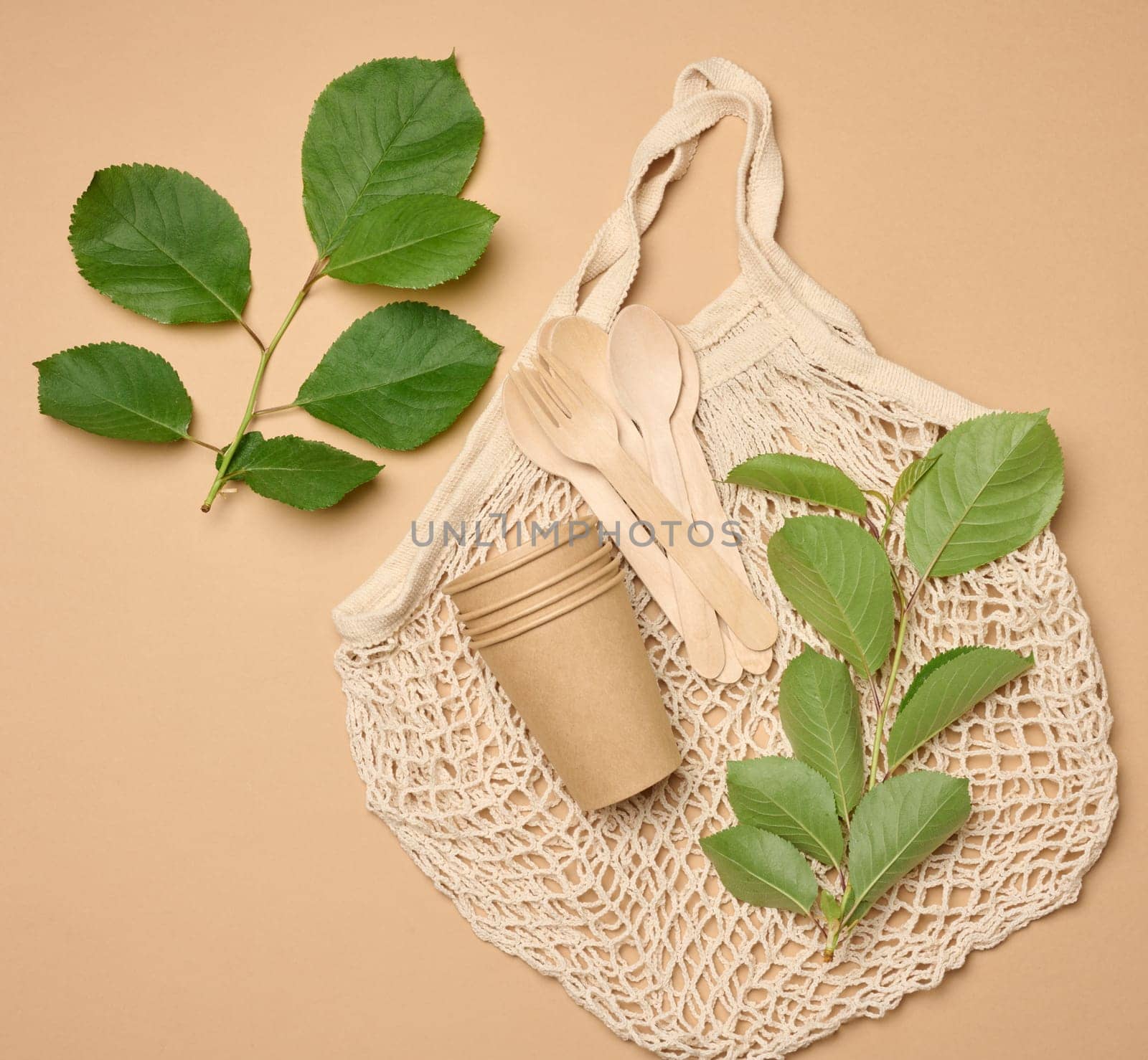 Textile knitted string bag and a stack of disposable cardboard glasses made of brown paper on a beige background and green leaves, top view