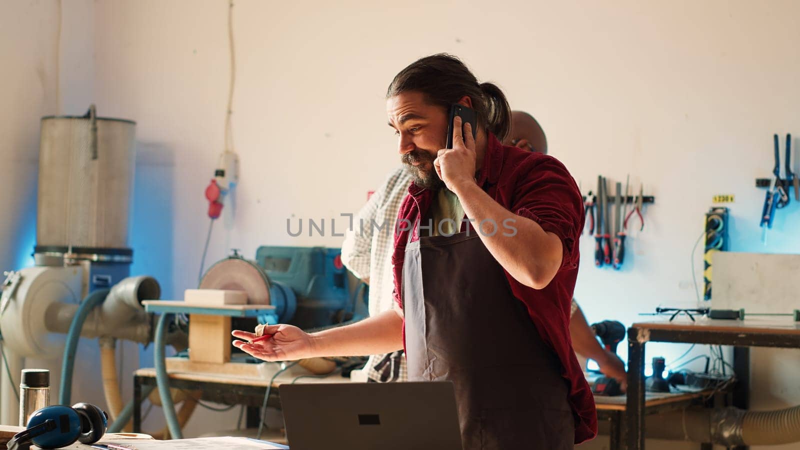 Manufacturer at work receiving telephone call from client inquiring about commission progress. Carpenter in assembly shop having friendly conversation on cellphone with customer, camera A