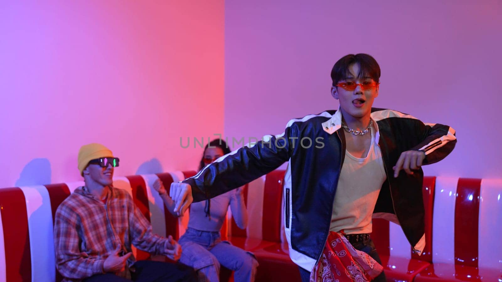 Asian man standing and dancing with pop music while multicultural friend sitting behind at home with led light. Attractive street dancer moving to hip hop music while looking at camera. Regalement.