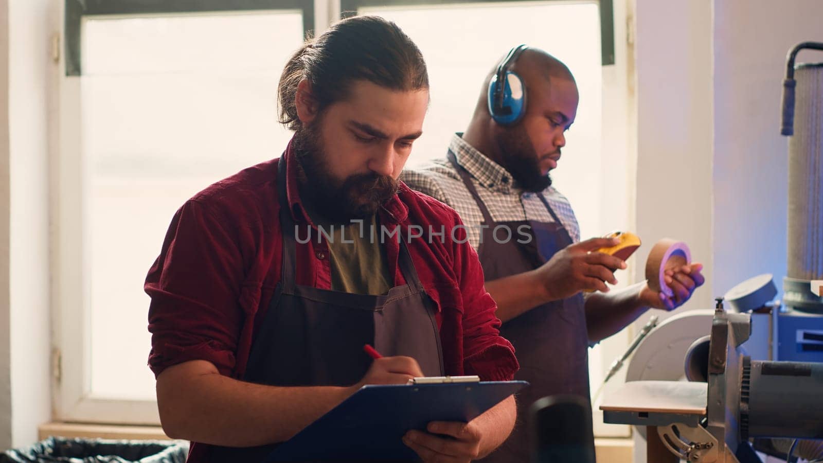 Craftsperson drawing blueprints on notepad in studio with BIPOC coworker in background solving tasks. Manufacturer looking at technical schematics to execute woodworking projects, camera A