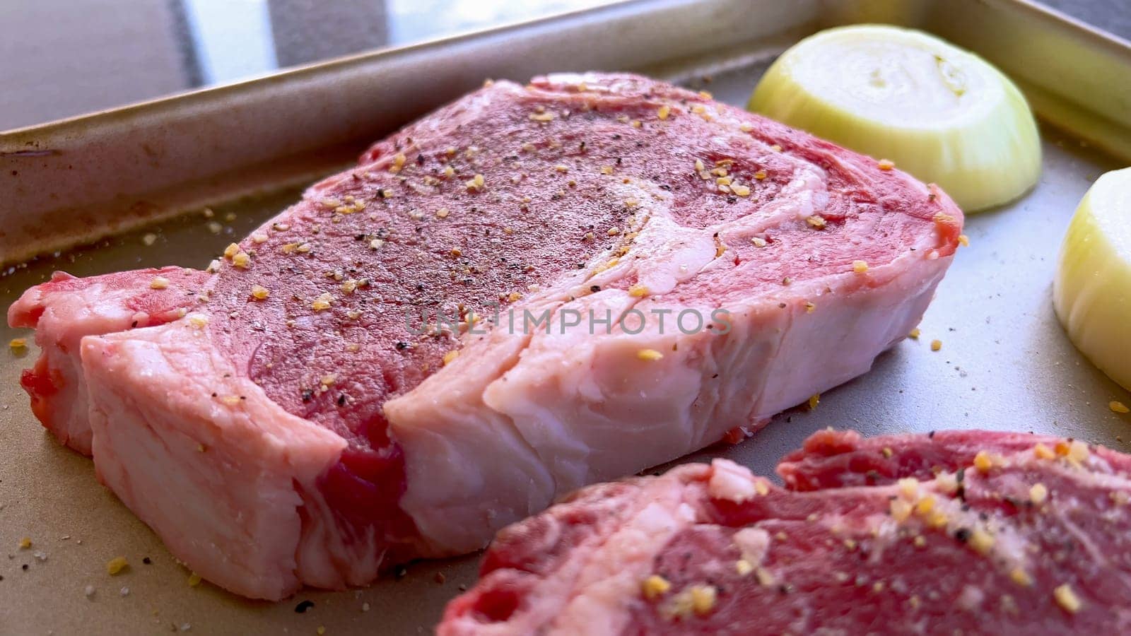 This image showcases three raw ribeye steaks, generously seasoned with coarse spices, alongside halves of fresh onions on a baking tray, prepared for a delicious grilling session.