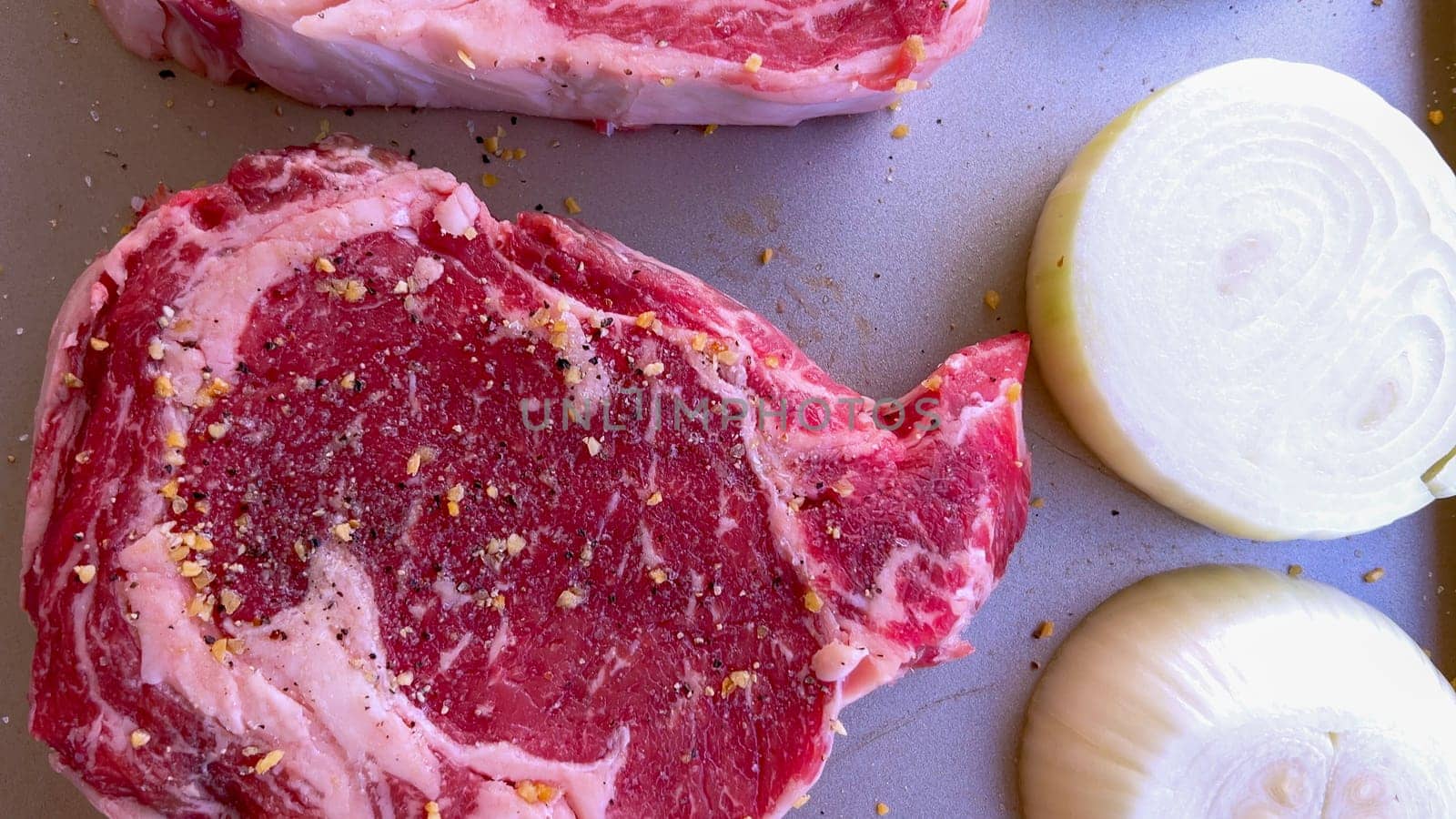 This image showcases three raw ribeye steaks, generously seasoned with coarse spices, alongside halves of fresh onions on a baking tray, prepared for a delicious grilling session.