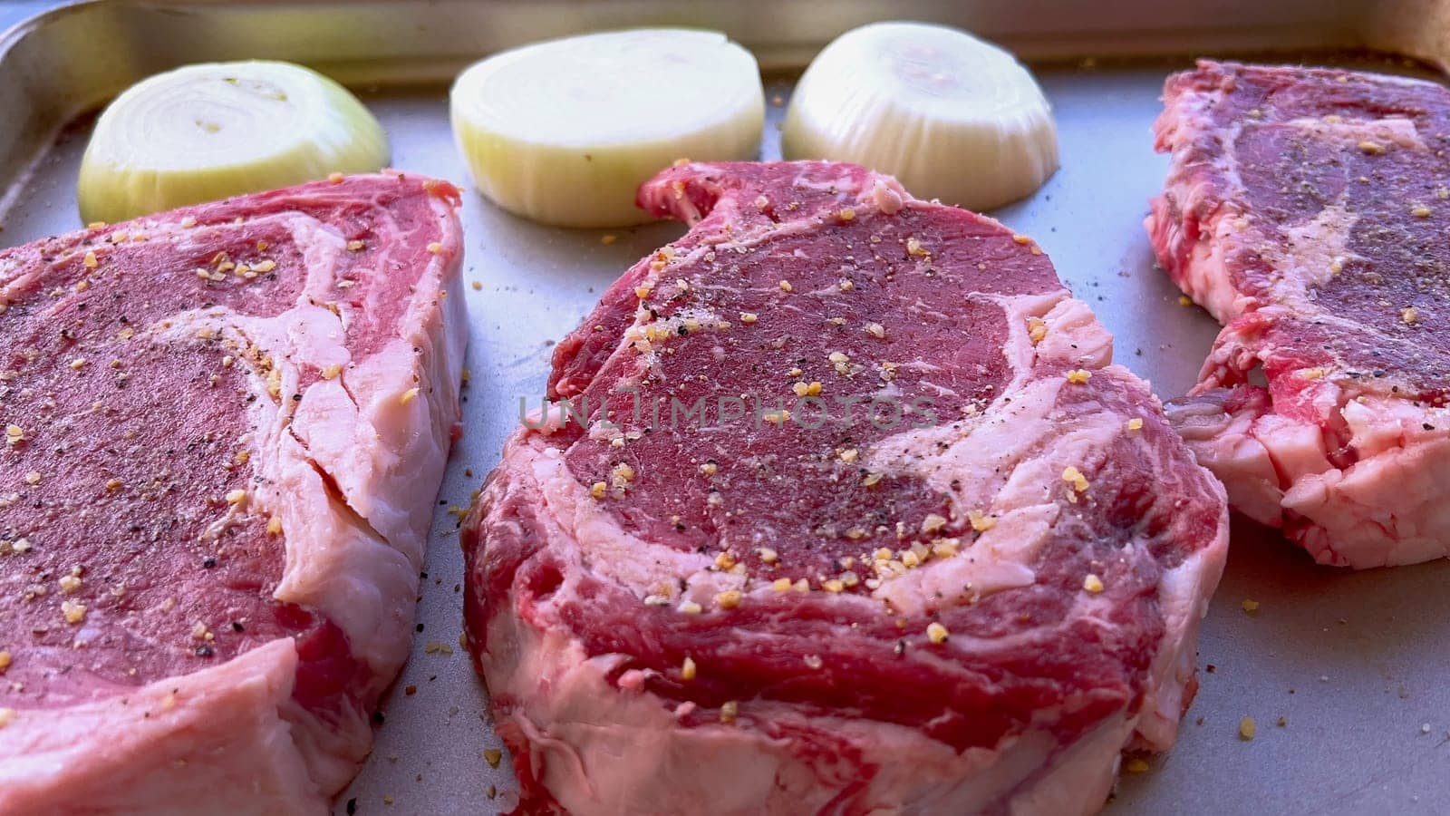 This image showcases three raw ribeye steaks, generously seasoned with coarse spices, alongside halves of fresh onions on a baking tray, prepared for a delicious grilling session.