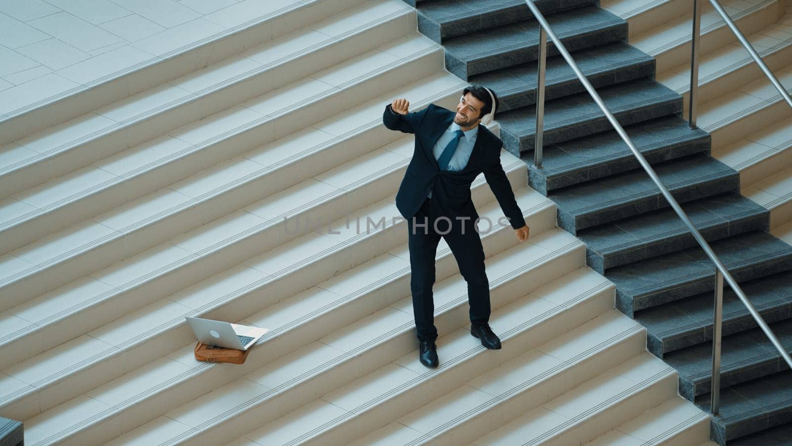Professional business man standing at stair while moving to music from headphone. Skilled project manager dancing while listening relax sound with laptop placed on stairs. Modern dance. Exultant.
