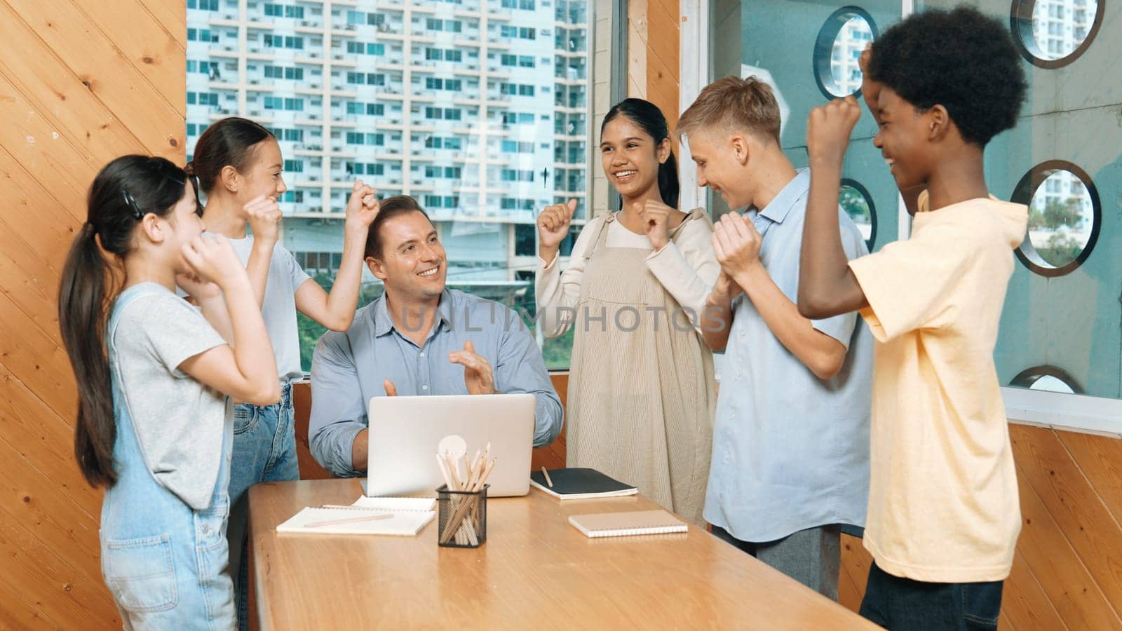 Caucasian teacher and multicultural students clapping hand or putting the hand in the air together to celebrate for successful project with laptop and equipment placed on table in class. Edification.