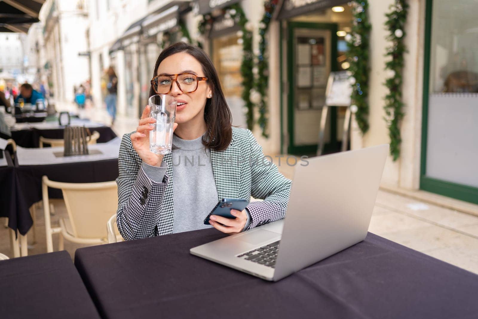 Portrait of attractive delighted charming smiling woman freelancer or student wearing glasses, working in cafe on laptop, holding mobile phone in hands, drinking water from glass.