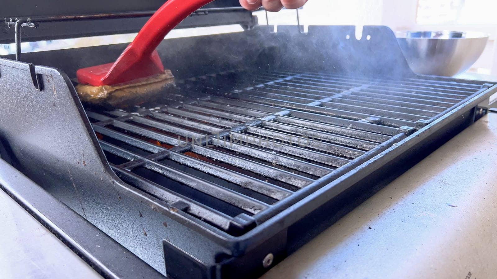 A hand uses a red grill brush to clean the black grates of a barbecue grill, ensuring it remains in perfect condition for the next grilling session.