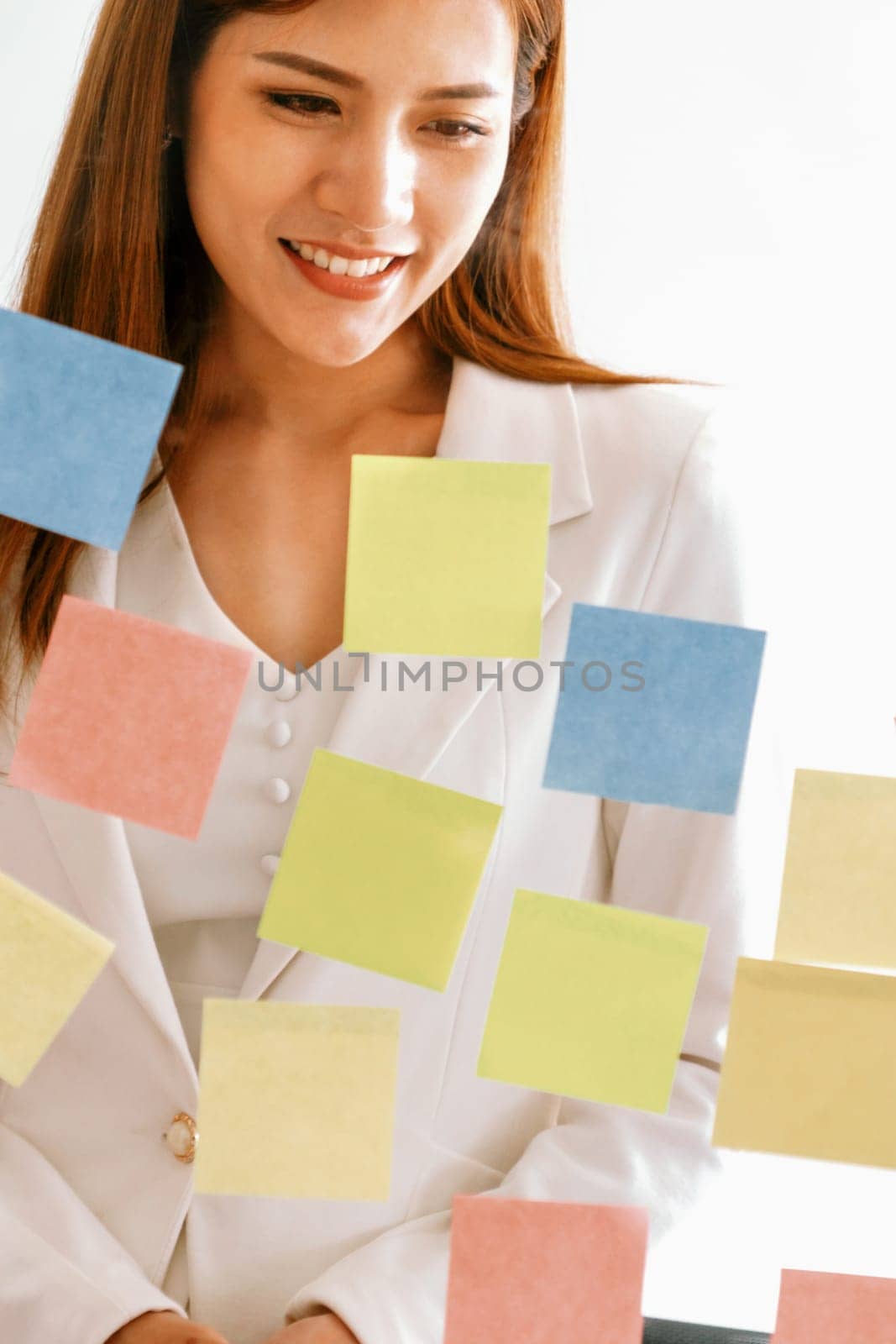 Happy businesswoman thinking creative ideas with sticky notes on glass wall at the office. Work planning and education concept uds