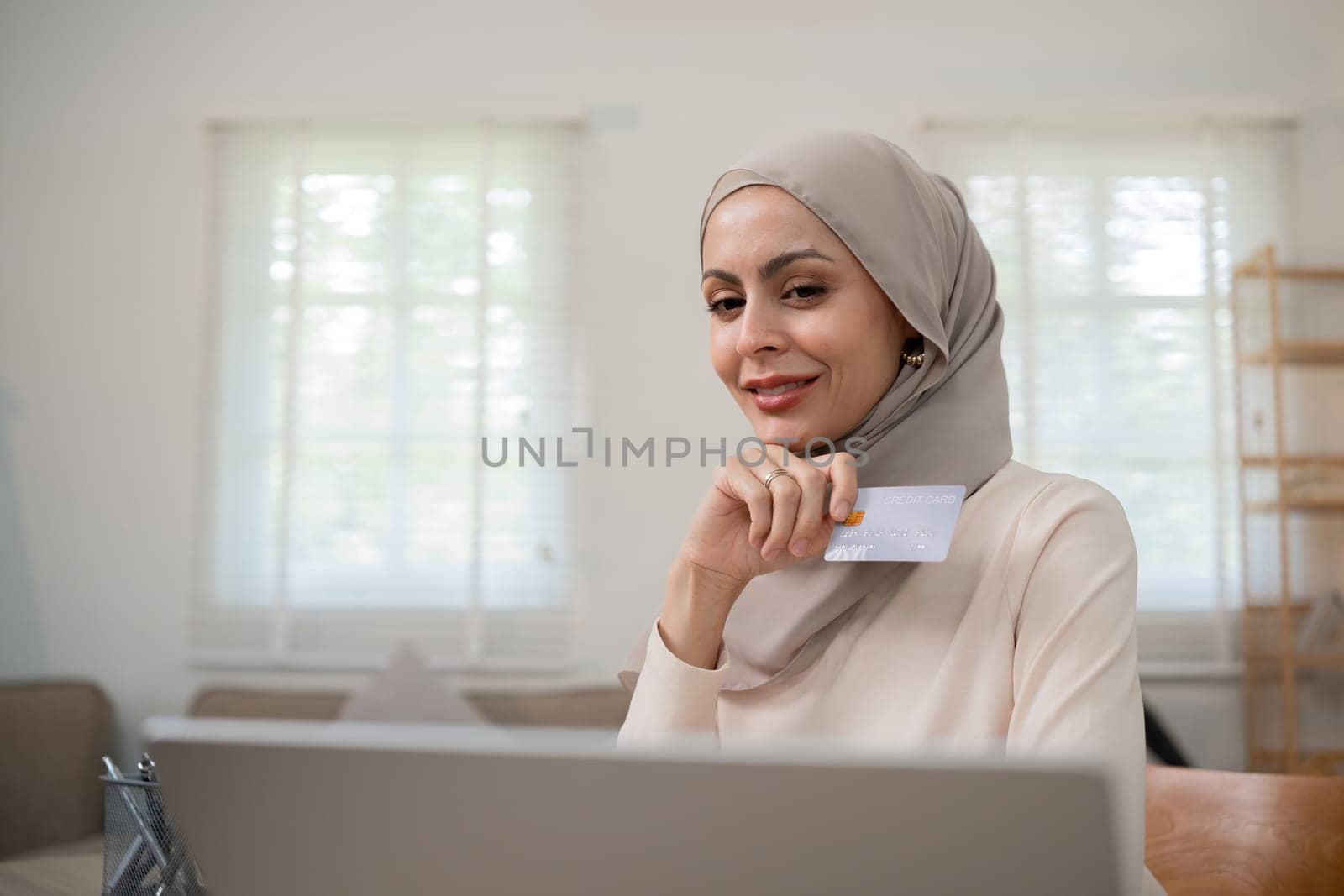 A young Muslim woman wearing a hijab sat contentedly shopping online and holding a credit card..