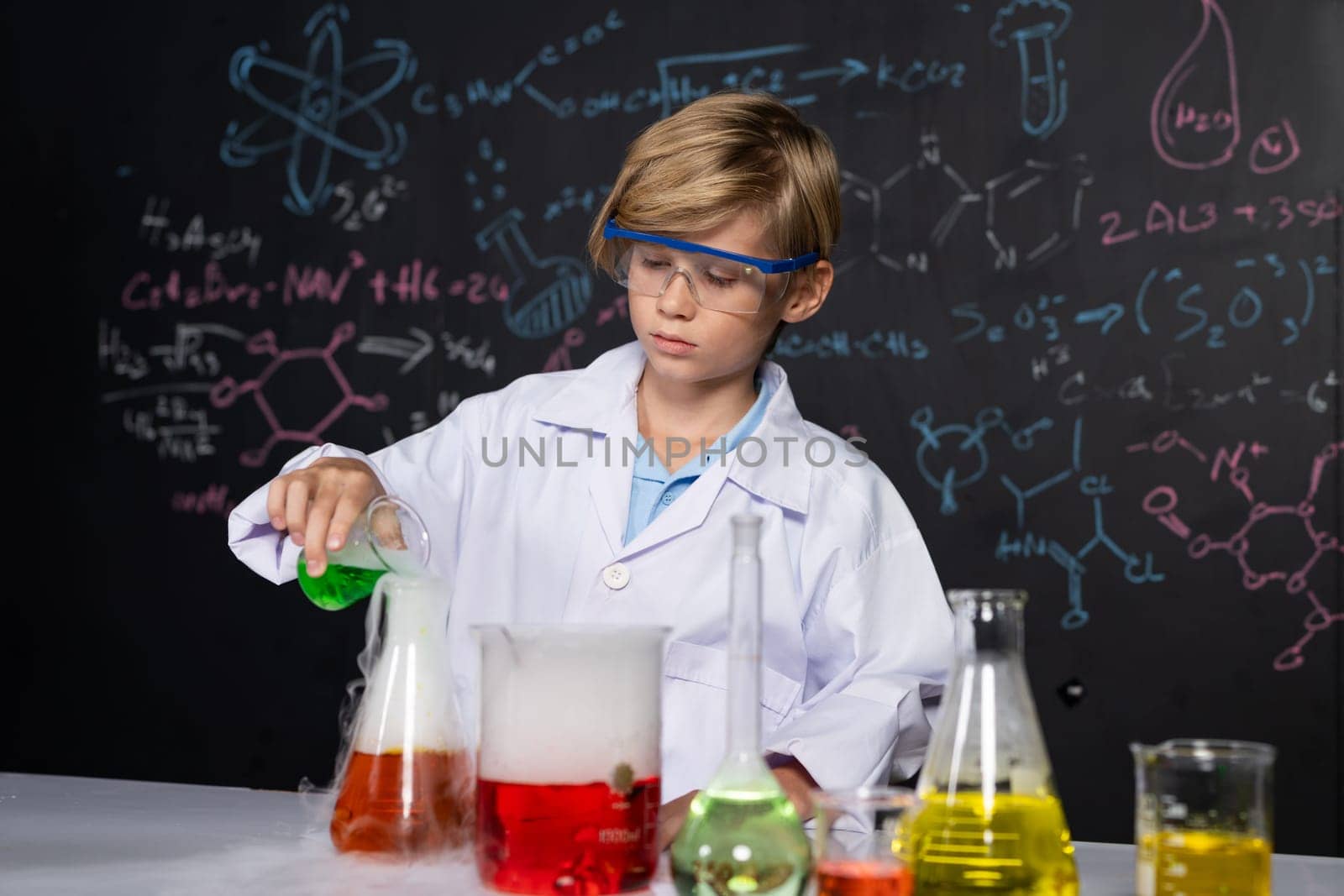 Blonde hair schoolboy in laboratory wear lab coat stand and learn science of chemistry technology in STEM class. The student pour liquid down to glass beaker. On the table put many flask. Erudition.