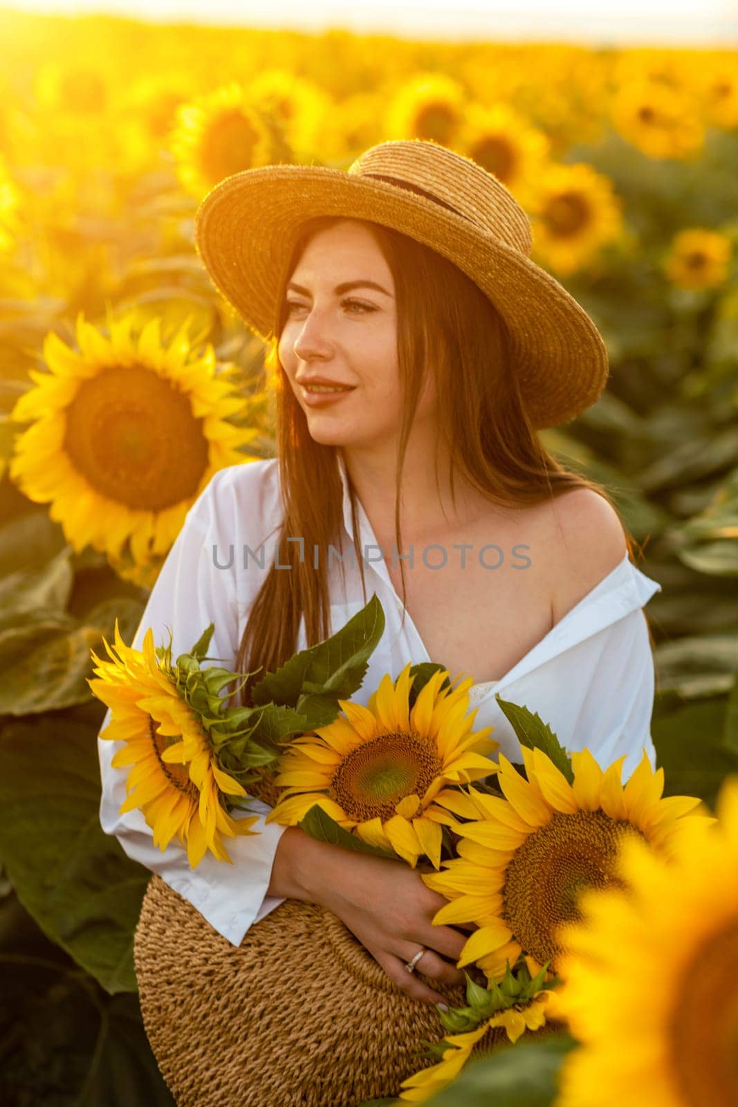 A girl in a hat on a beautiful field of sunflowers against the sky in the evening light of a summer sunset. Sunbeams through the flower field. Natural background