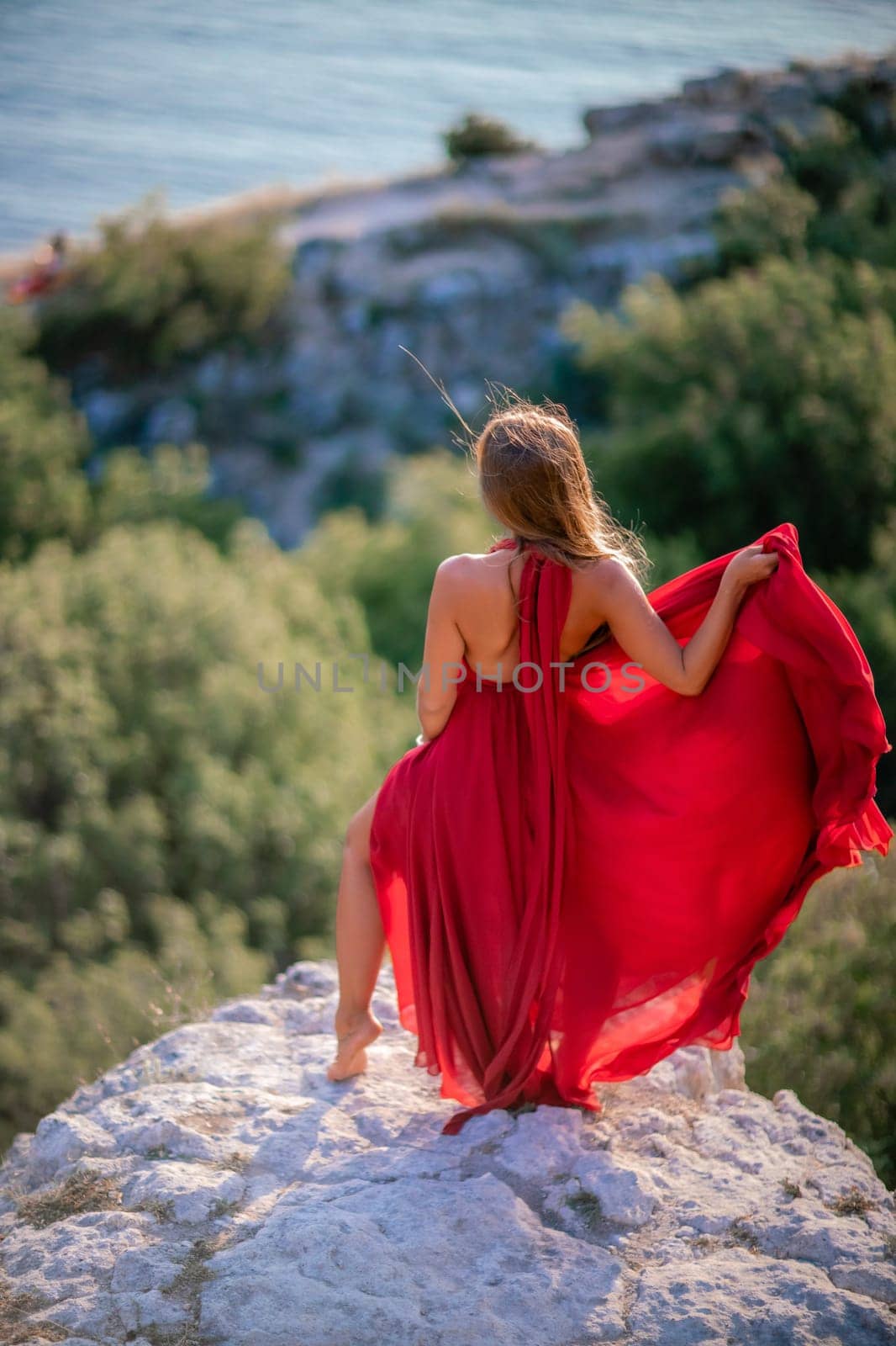 Woman sunset sea red dress, back view a happy beautiful sensual woman in a red long dress posing on a rock high above the sea on sunset