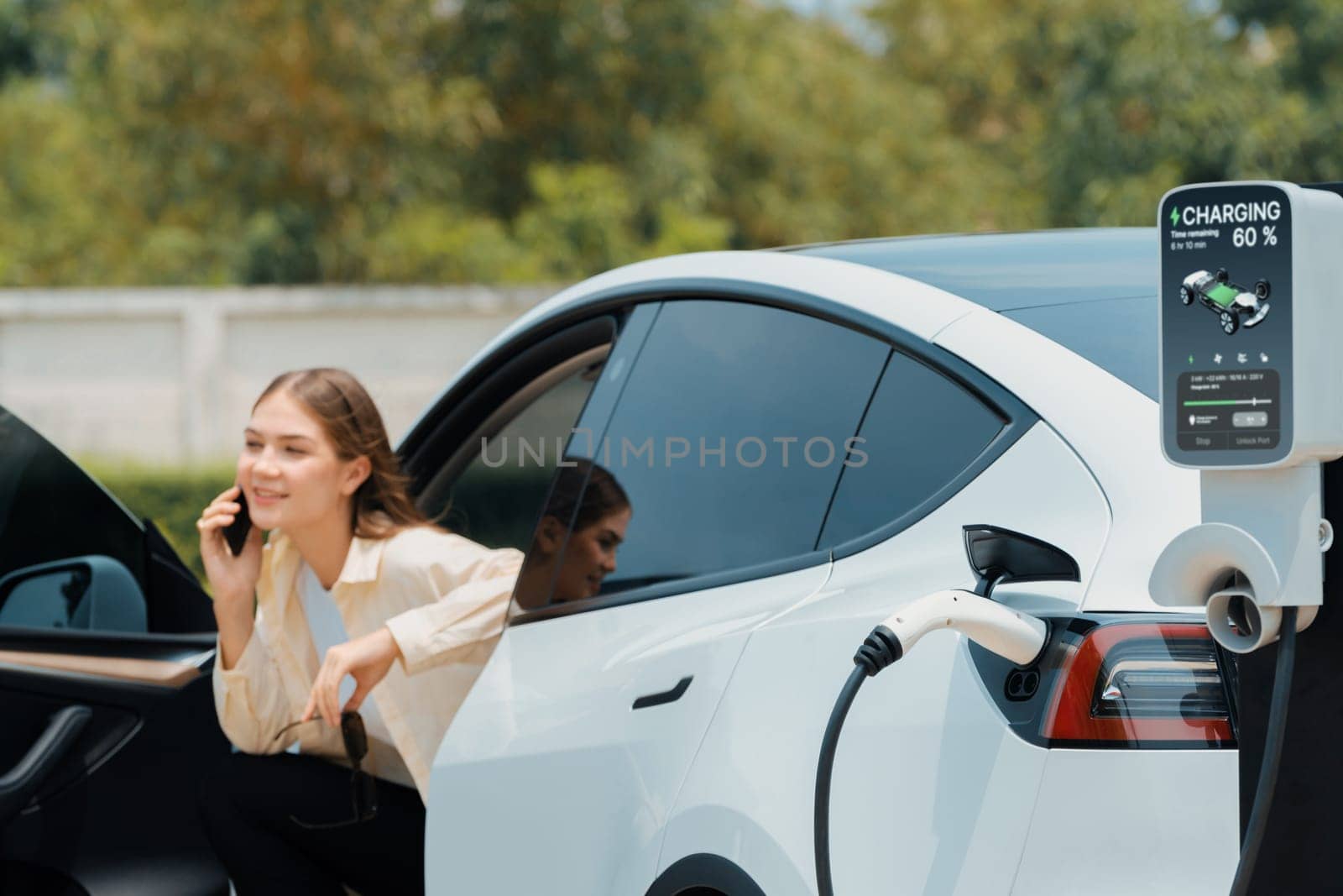 Young woman recharge her EV electric vehicle at green city park parking lot while talking on phone. Sustainable urban lifestyle for environment friendly EV car with battery charging station. Expedient