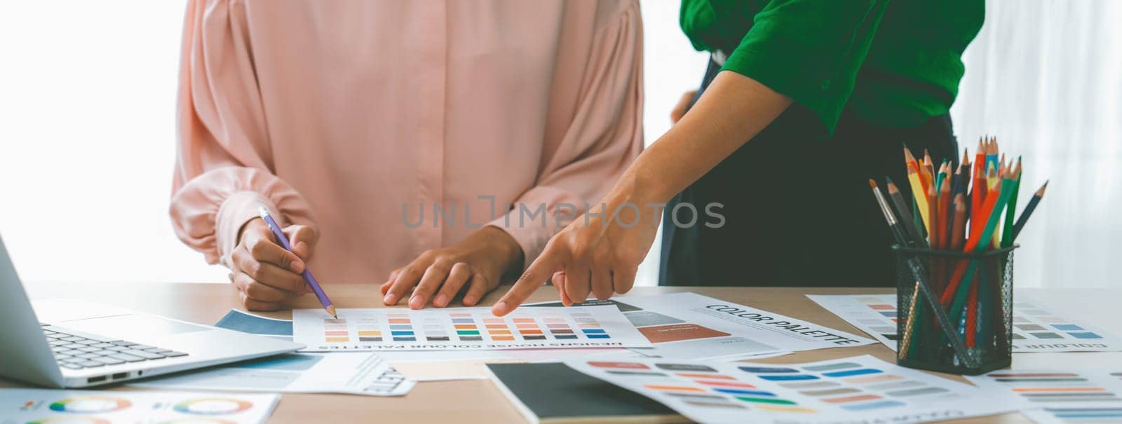 Cropped image of two female young beautiful graphic designer choose the color from color palate by using laptop on table with work tools and equipments scatter around at modern office. Variegated.