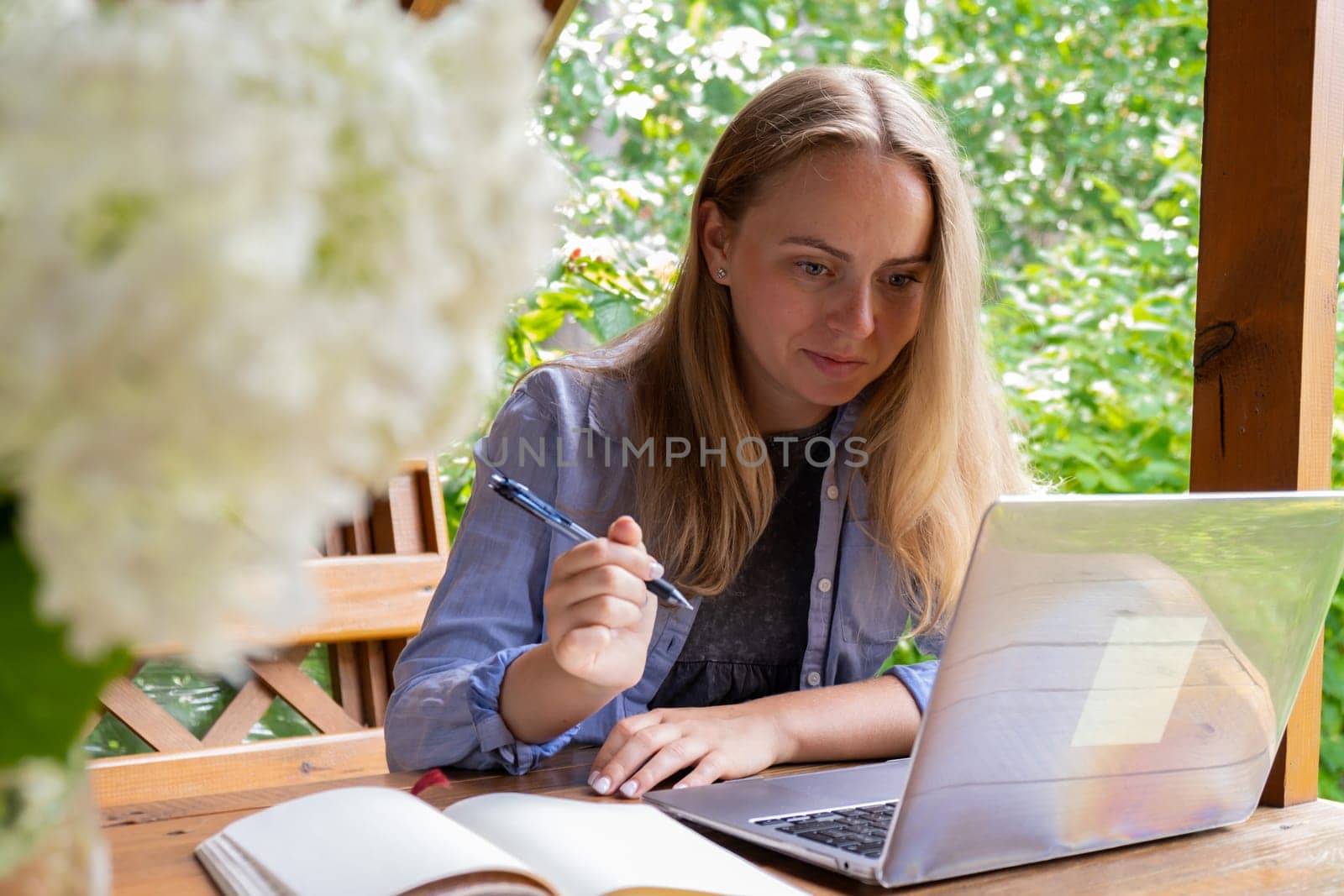 Female student has online lesson education outdoor in garden wooden alcove. Blonde woman sitting outside work on laptop having video call by anna_stasiia