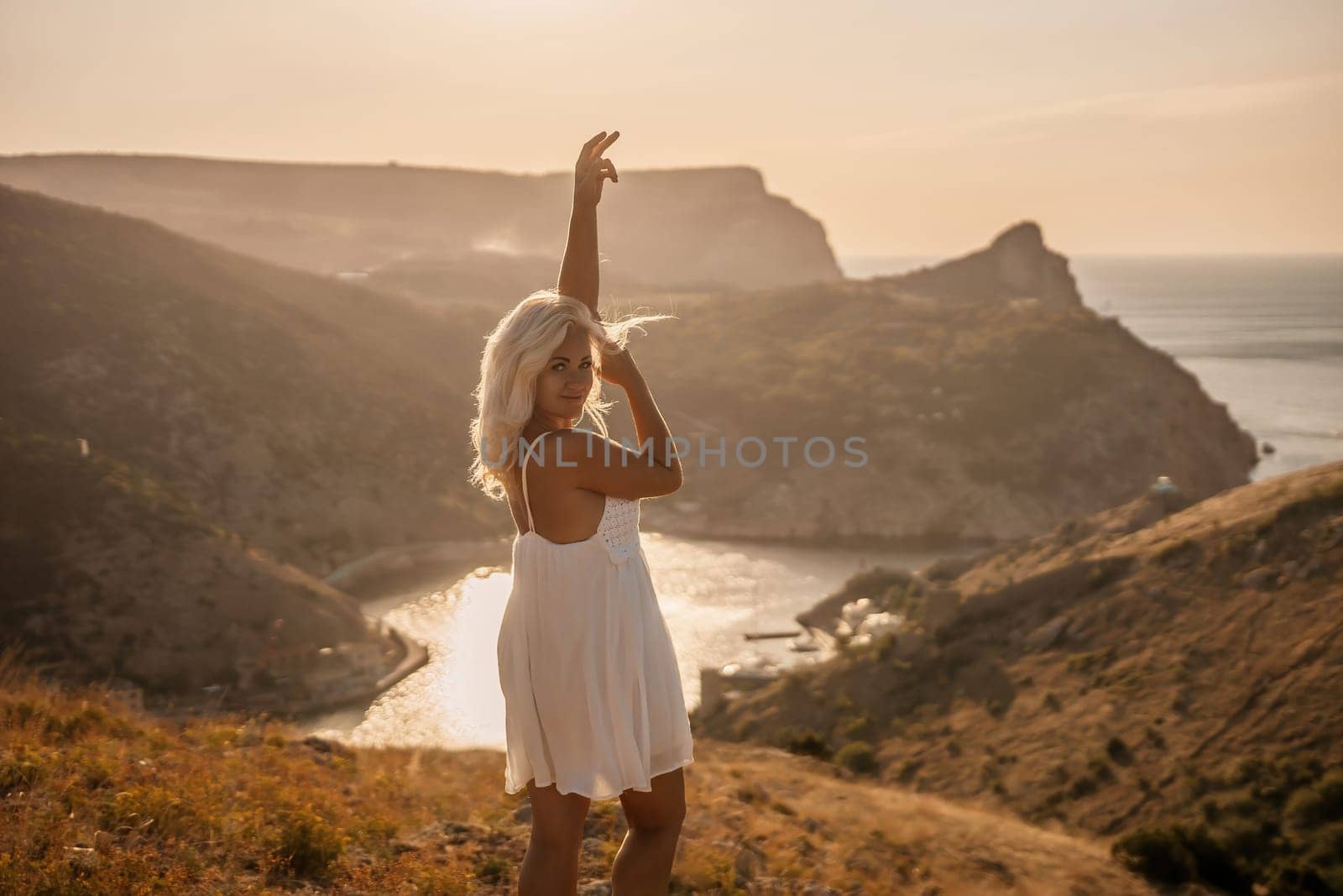 A woman stands on a hill overlooking a body of water. She is wearing a white dress and she is enjoying the view. by Matiunina