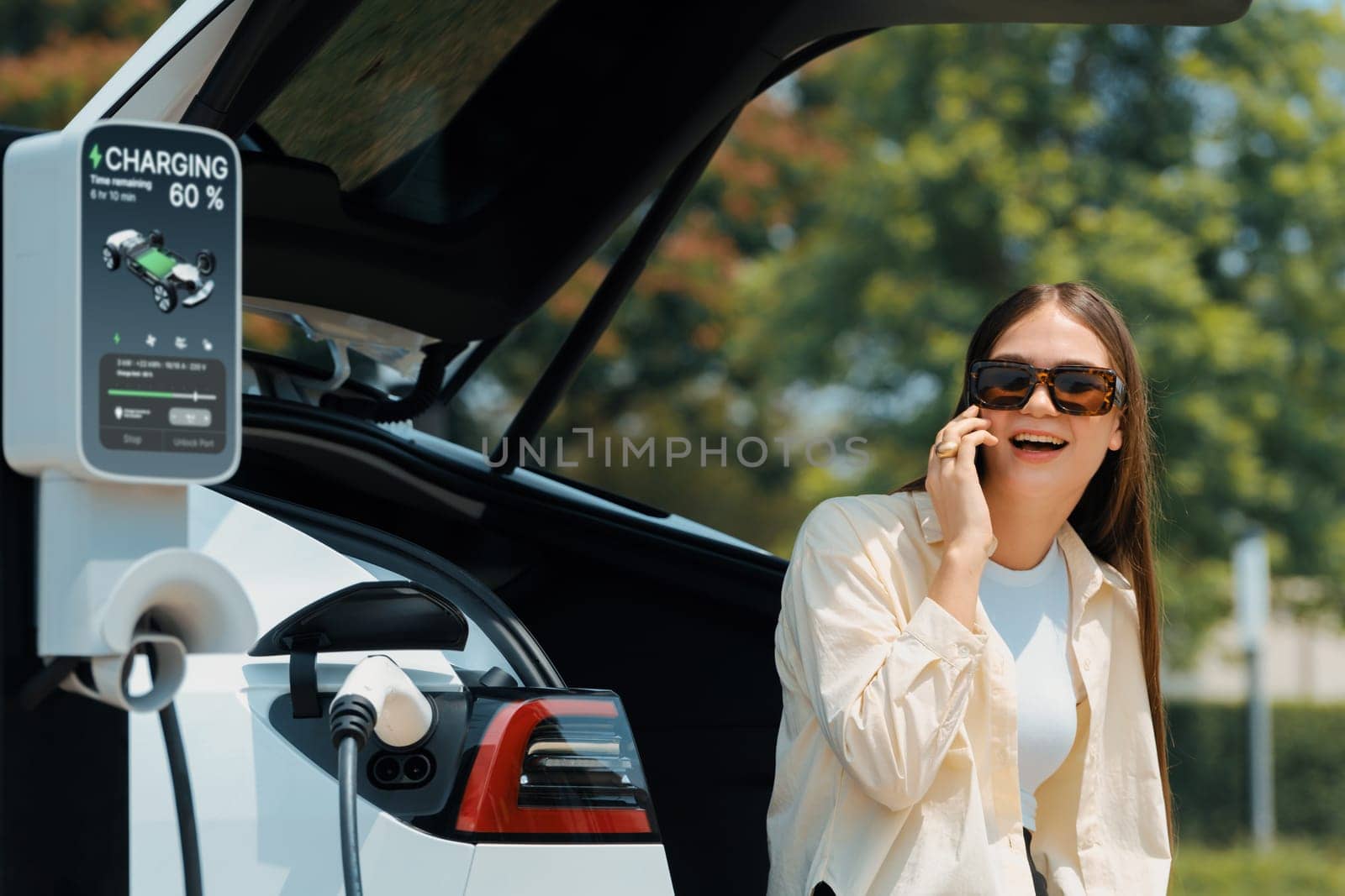 Young woman recharge her EV electric vehicle at green city park parking lot while talking on phone. Sustainable urban lifestyle for environment friendly EV car with battery charging station. Expedient