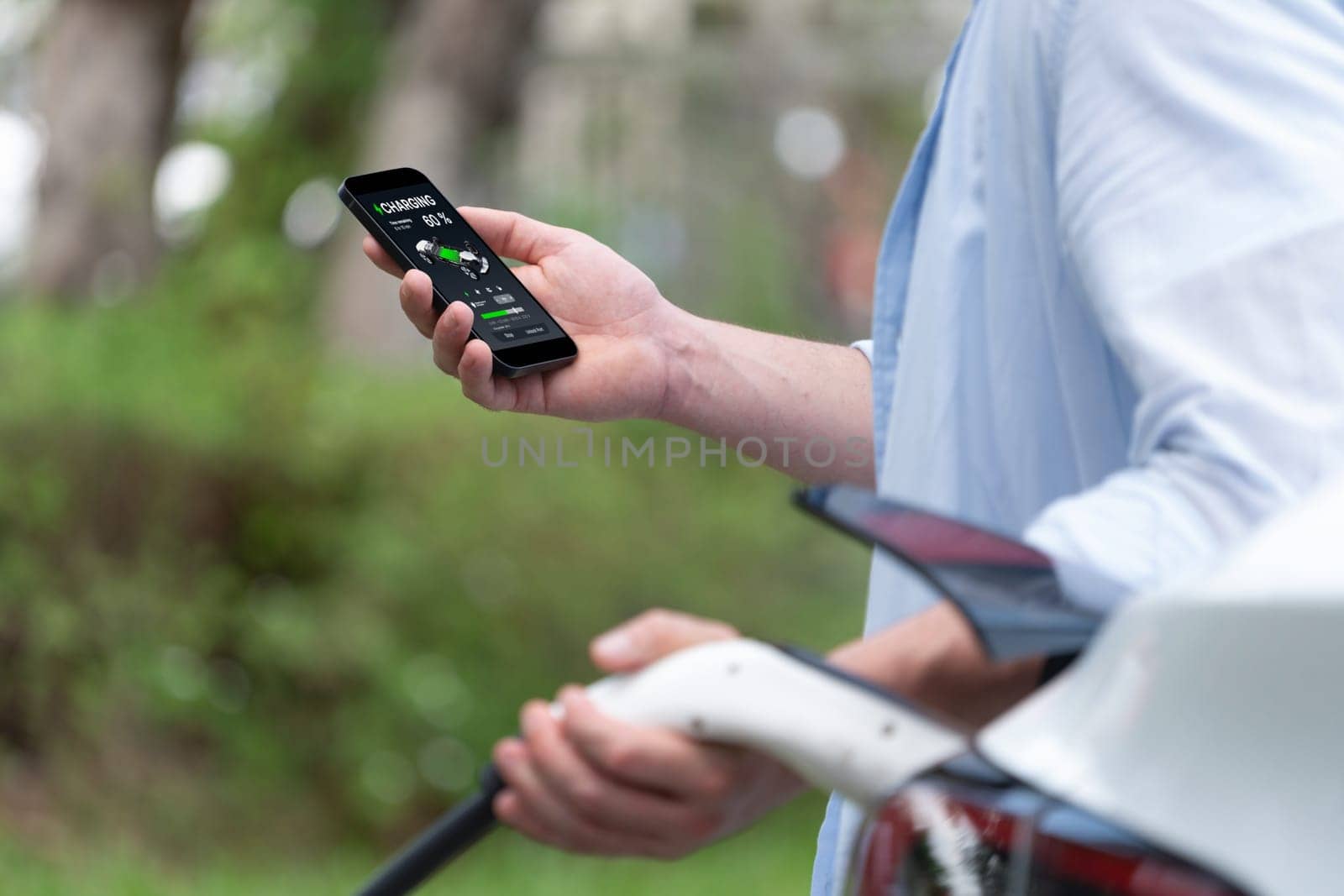 Modern eco-friendly man recharging electric vehicle from EV charging station, using Innovative EV technology utilization for tracking energy usage to optimize battery charging on smartphone.Synchronos