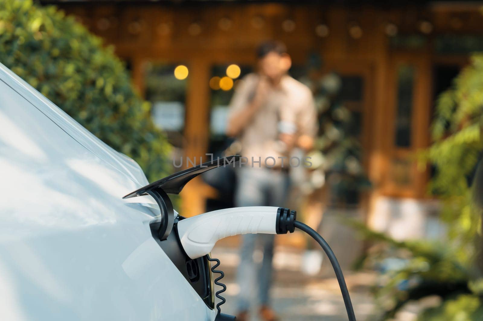 Focused EV electric car recharging at outdoor coffee cafe in springtime garden with blur background of eco friendly man, green city sustainability and environmental friendly EV vehicle. Expedient