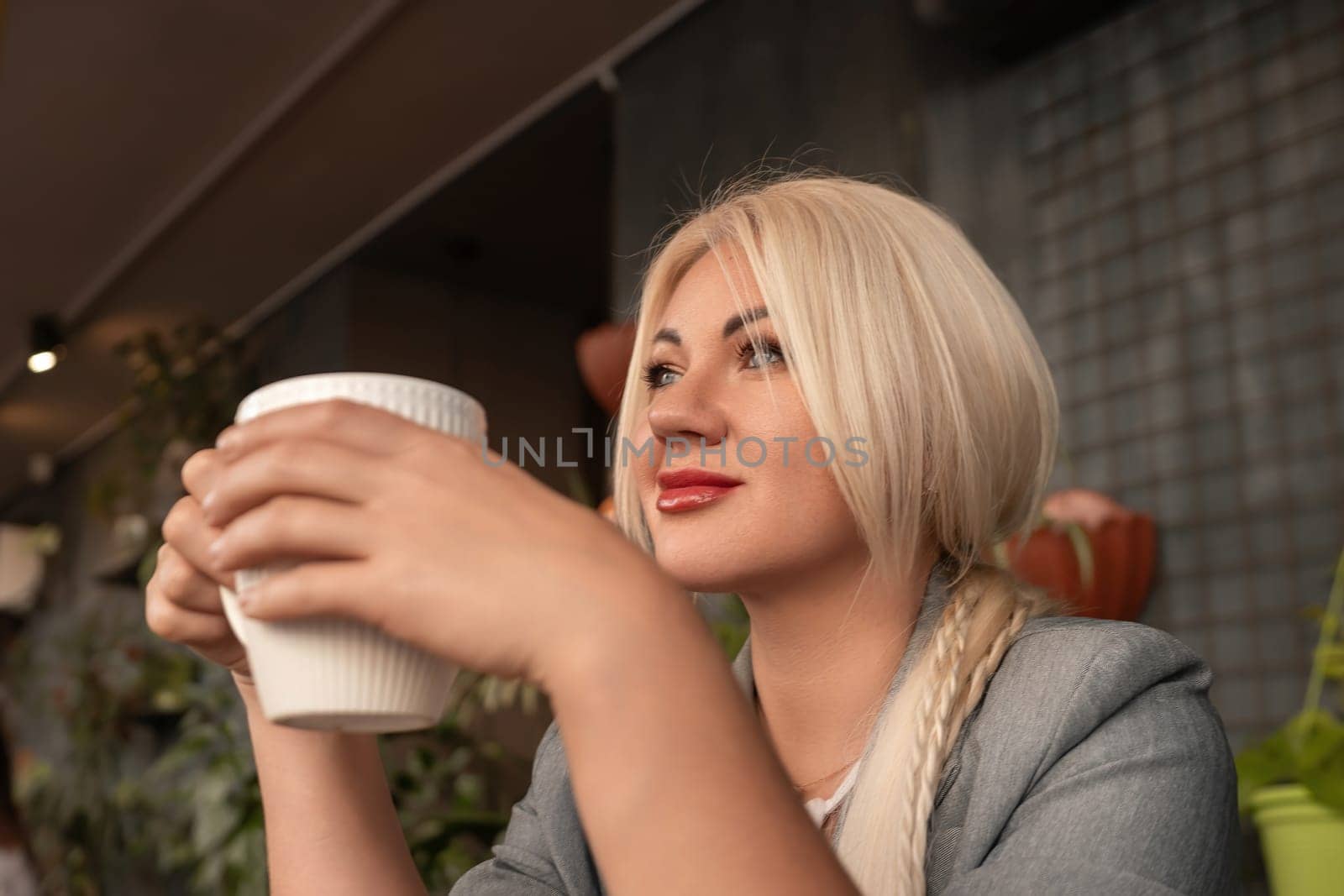 A blonde woman is holding a white coffee cup and looking at something. She is wearing a blue jacket and has red lipstick