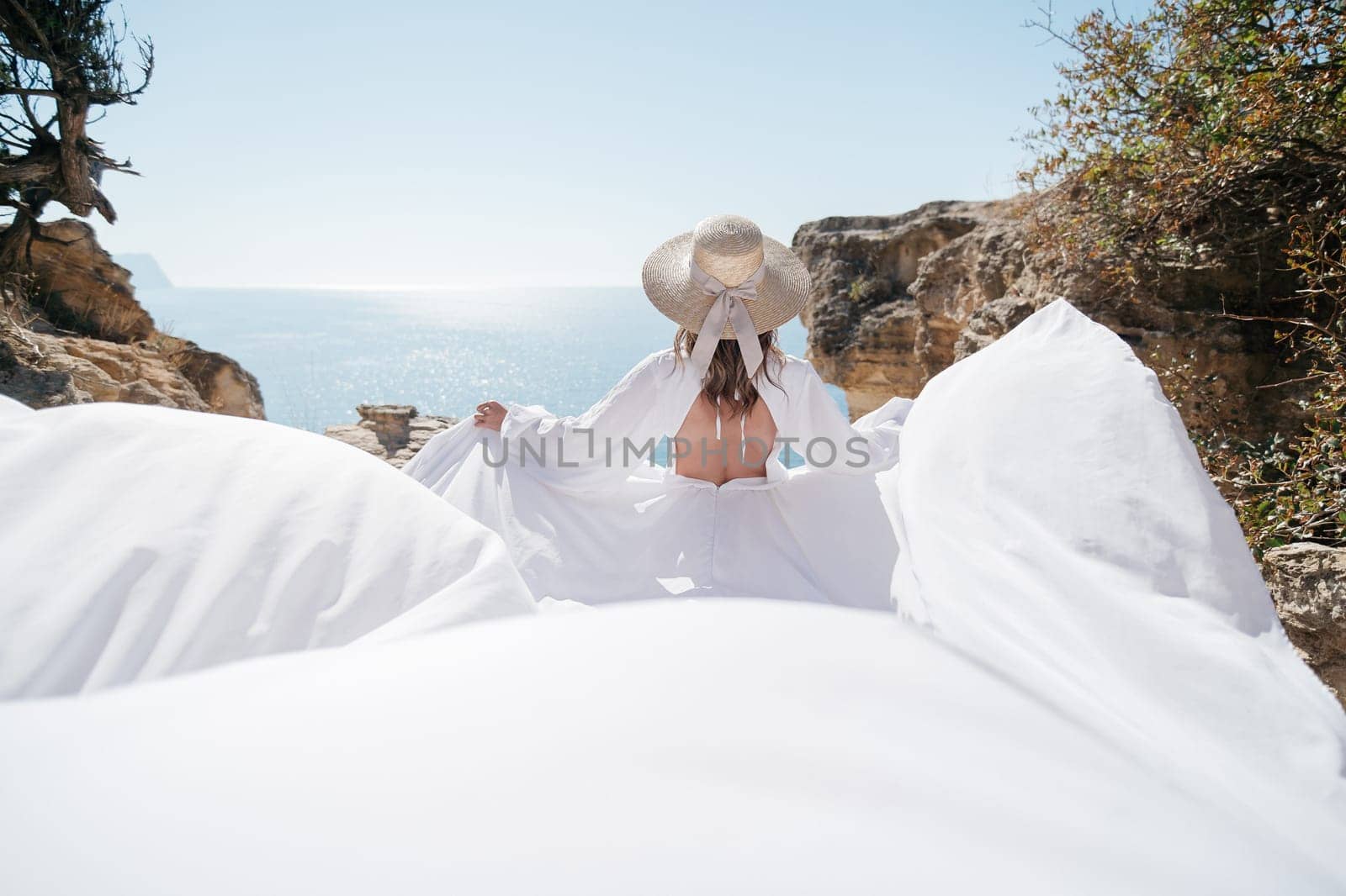 A woman in a white dress is standing on a rocky beach with her hat on. The scene is serene and peaceful, with the woman enjoying the view of the water by Matiunina