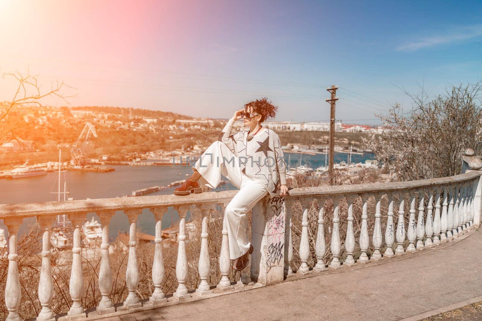 Woman walks around the city, lifestyle. A young beautiful woman in white trousers and a sweater sits on a white fence with balusters and overlooks the sea bay and the city. by Matiunina