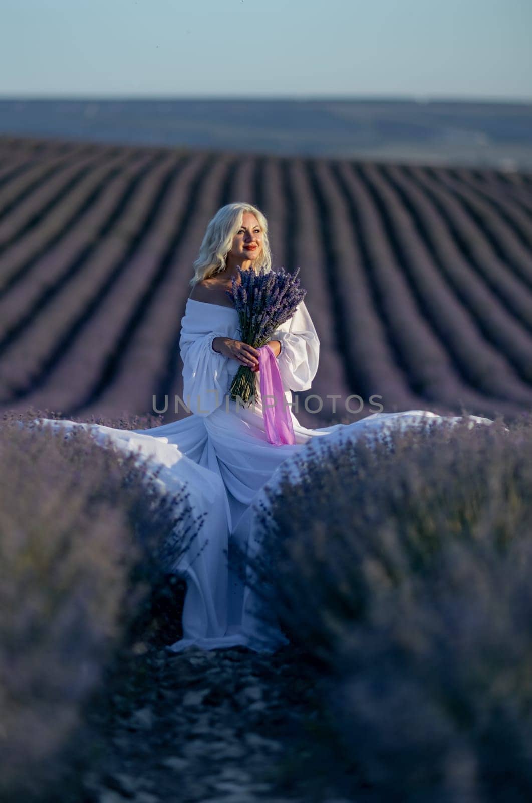 Blonde woman poses in lavender field at sunset. Happy woman in white dress holds lavender bouquet. Aromatherapy concept, lavender oil, photo session in lavender by Matiunina