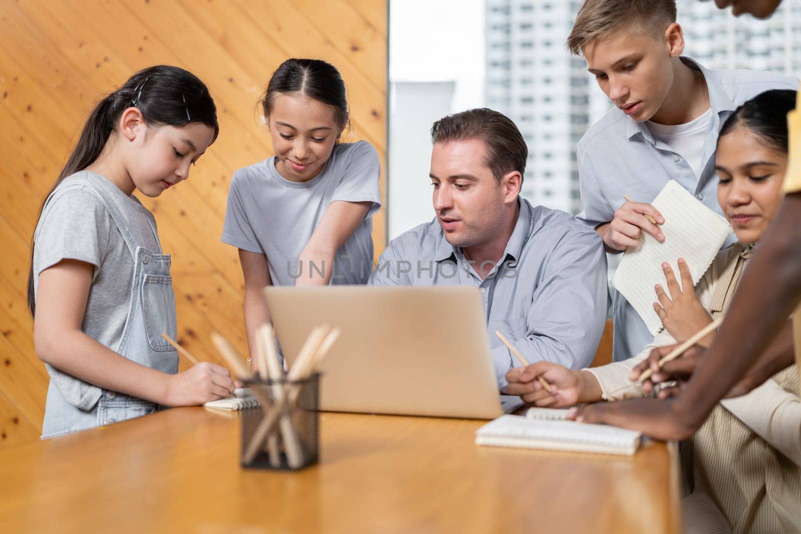 During a lecture course teacher sitting and use laptop. Students standing and separate to two group for taking note together using pencil also talk about lesson. On table put pencil box. Edification.