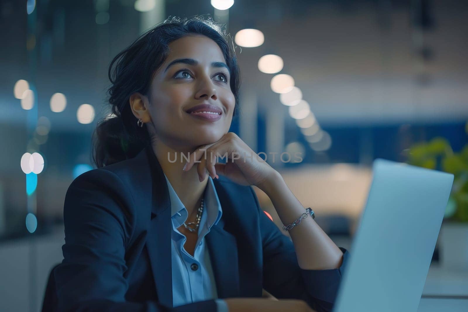 A woman in a business suit is sitting at a desk with a laptop in front of her. She is smiling and looking at the screen, possibly working or browsing the internet. Concept of productivity and focus