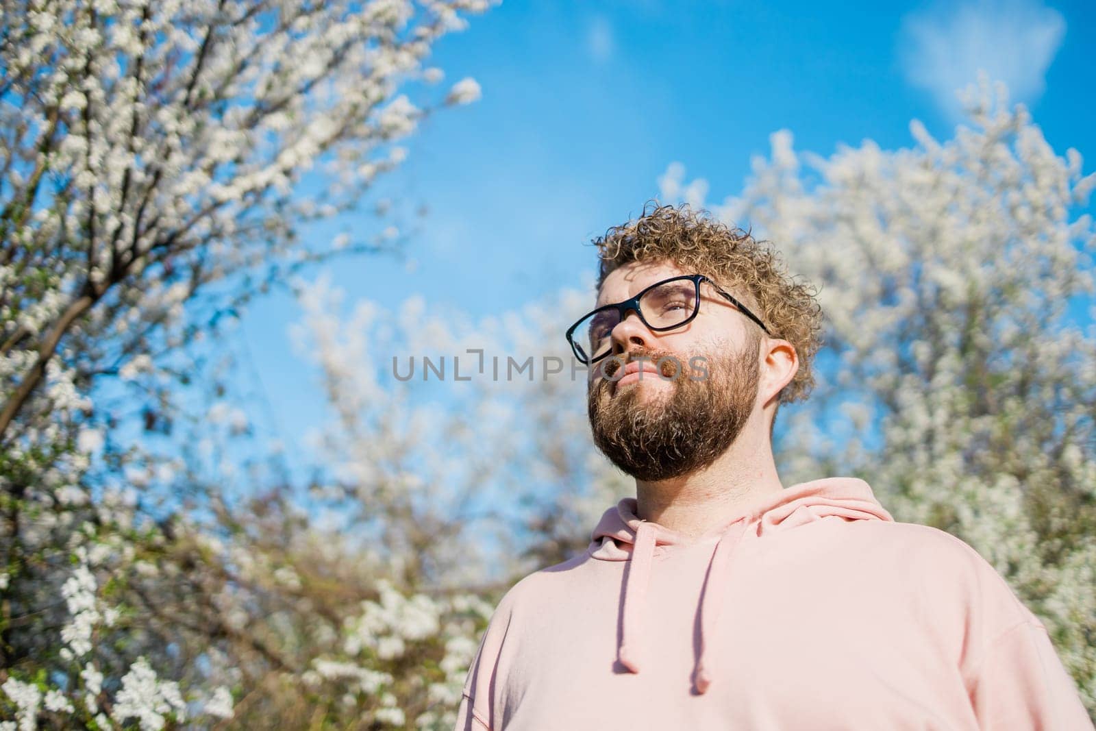 Male bearded guy standing under branches with flowers of blooming almond or cherry tree in spring garden. Spring blossom. Copy space