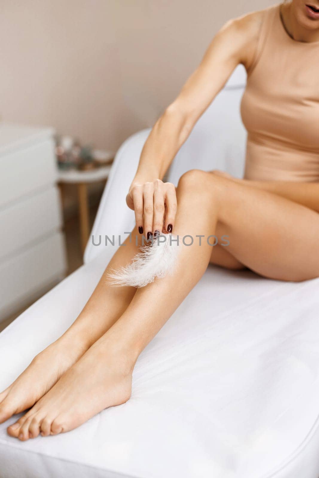 Woman touches her legs with white feather. Concepts of skin care and hair removal treatment. Female feet with smooth skin and soft ostrich feathe.